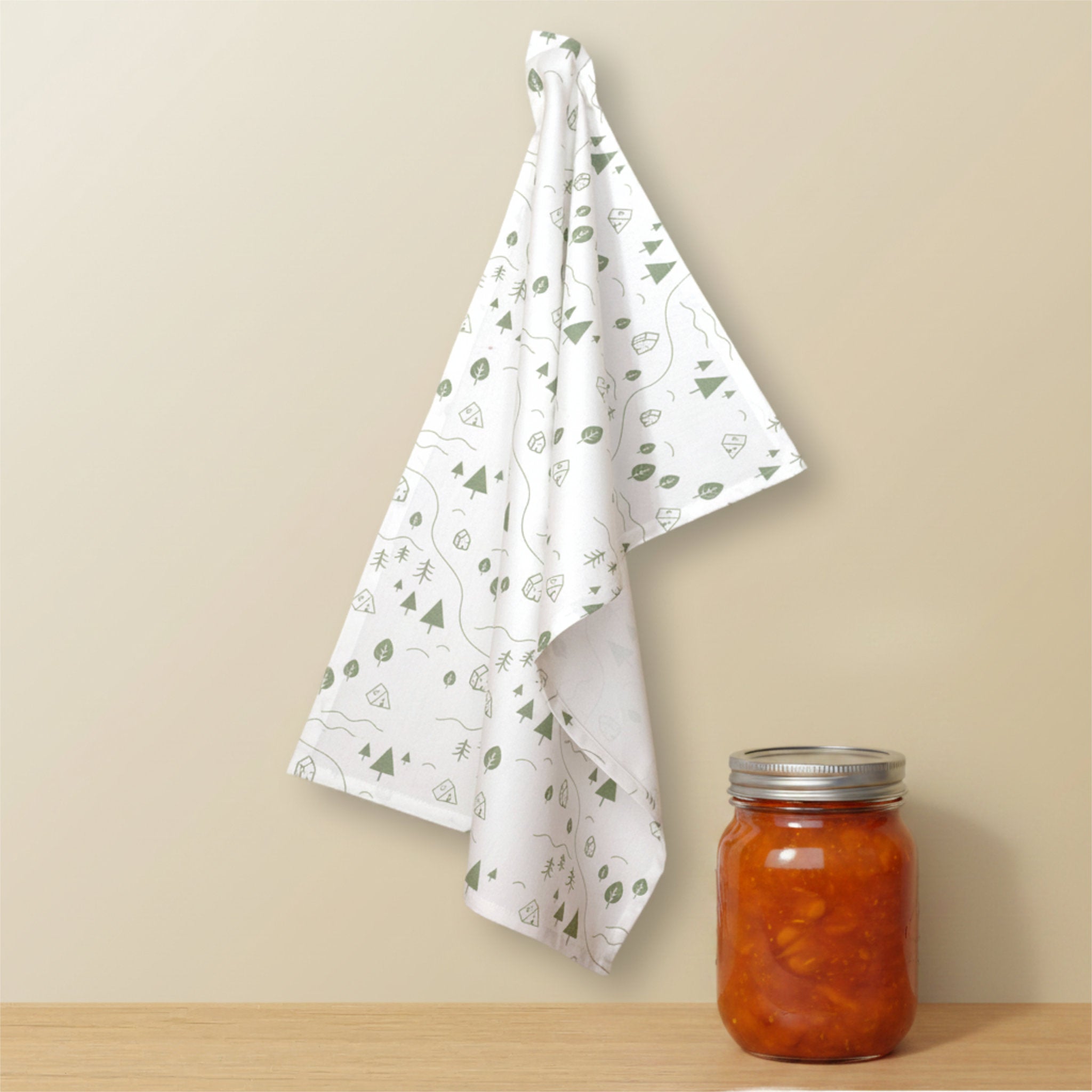 A white kitchen tea towel with a green minimalist tents and tree pattern hangs on a beige wall above a wooden surface, next to a glass mason jar filled with orange-red pickled fruit from Hem And Tassel
