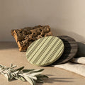Two round heatsink style cork trivets with striped grooves pattern in sage green and dark gray, styled on a beige surface with cork bark, a linen cloth, and dried leaves from Hem And Tassel 