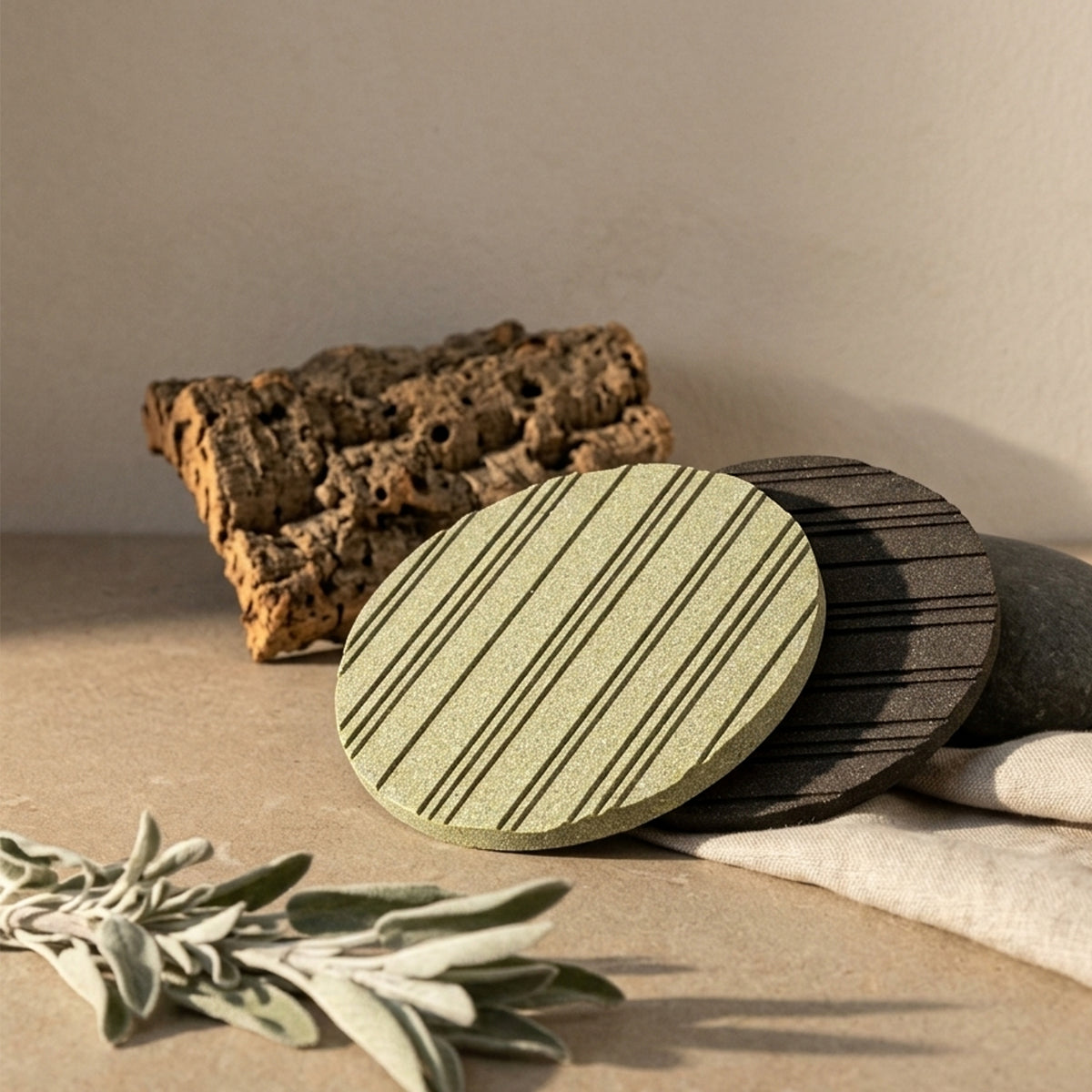 Two round heatsink style cork trivets with striped grooves pattern in sage green and dark gray, styled on a beige surface with cork bark, a linen cloth, and dried leaves from Hem And Tassel 