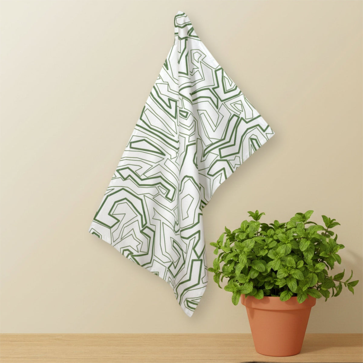 Hem And Tassel Squiggles cotton kitchen towel with green geometric pattern hanging on a wall near a potted plant.