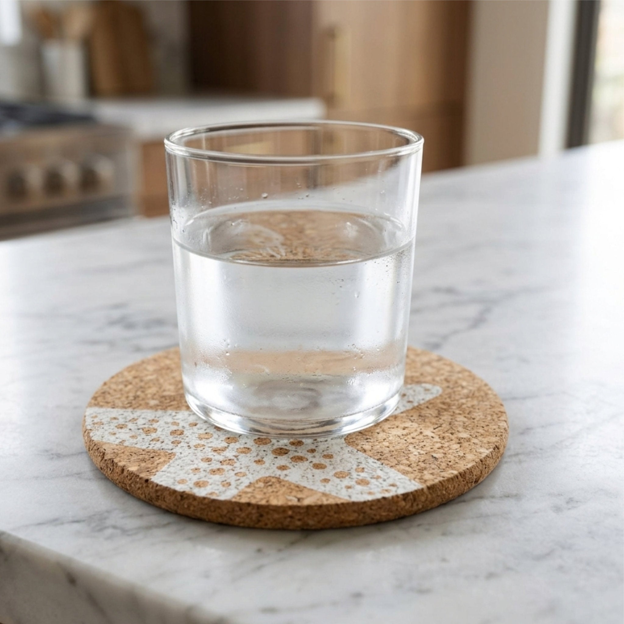 A clear glass tumbler filled with water sits on a round cork home by the sea coaster with a white starfish pattern, placed on a white marble countertop in a kitchen setting from Hem And Tassel 