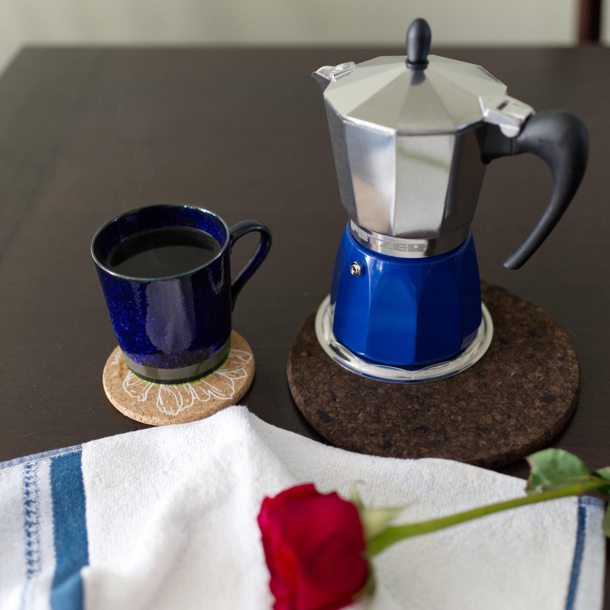 Blue espresso induction moka pot with a blue mug on a dark surface, accompanied by a red rose and a striped towel. from Hem And Tassel 