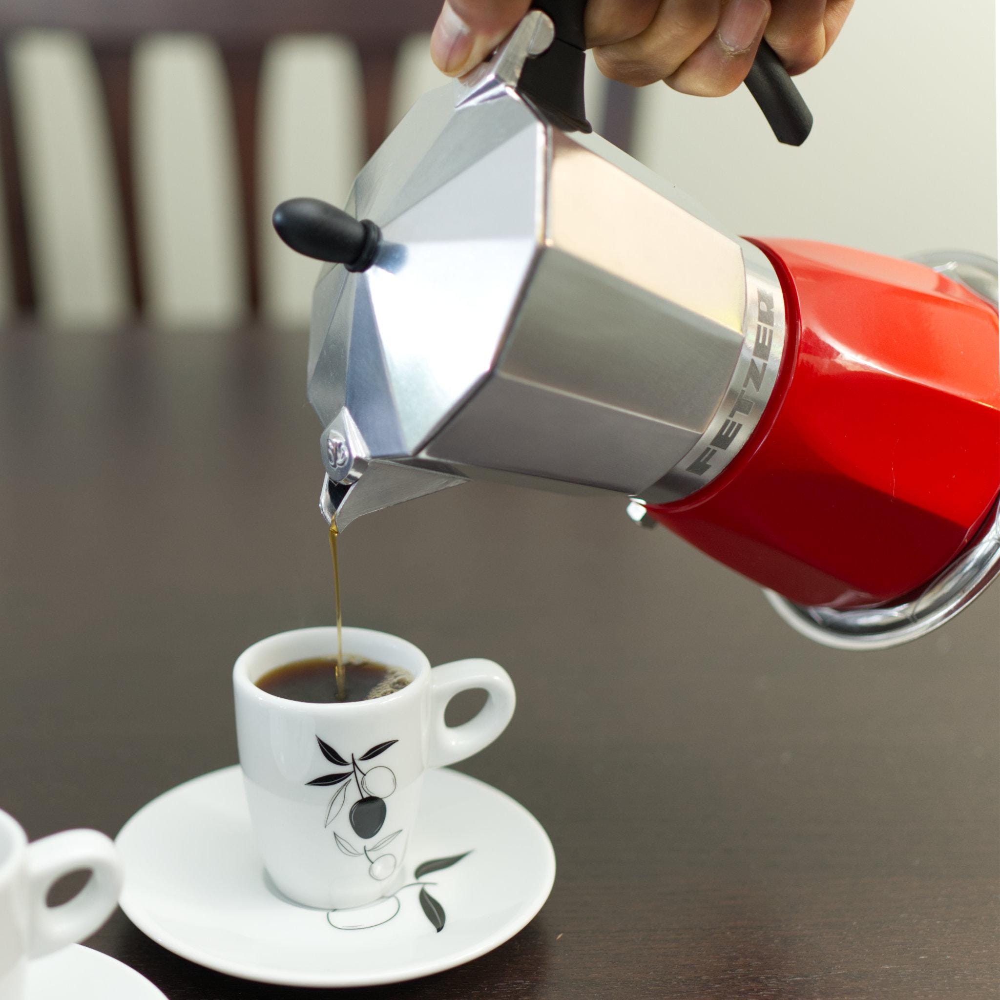 Red and silver induction Moka pot pouring coffee into a white cup with black design on a wooden table from Hem And Tassel 