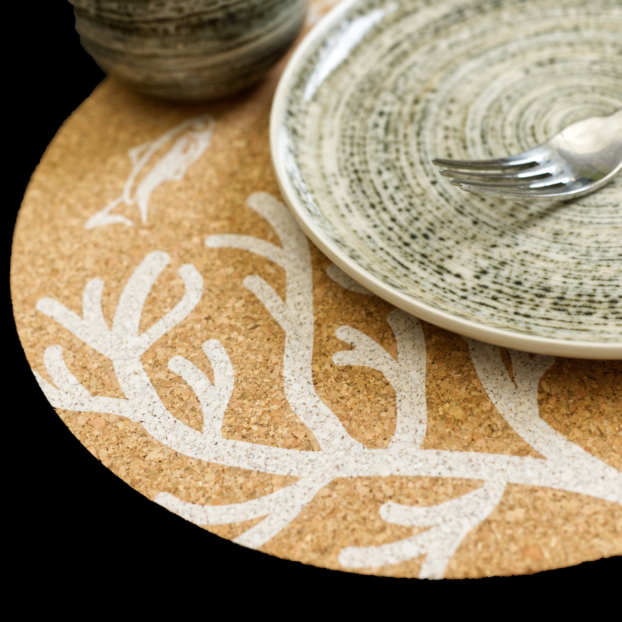 A round home by the sea cork placemat with a white printed fish and coral pattern, styled with a speckled ceramic plate and a silver fork on a black background from Hem And Tassel 