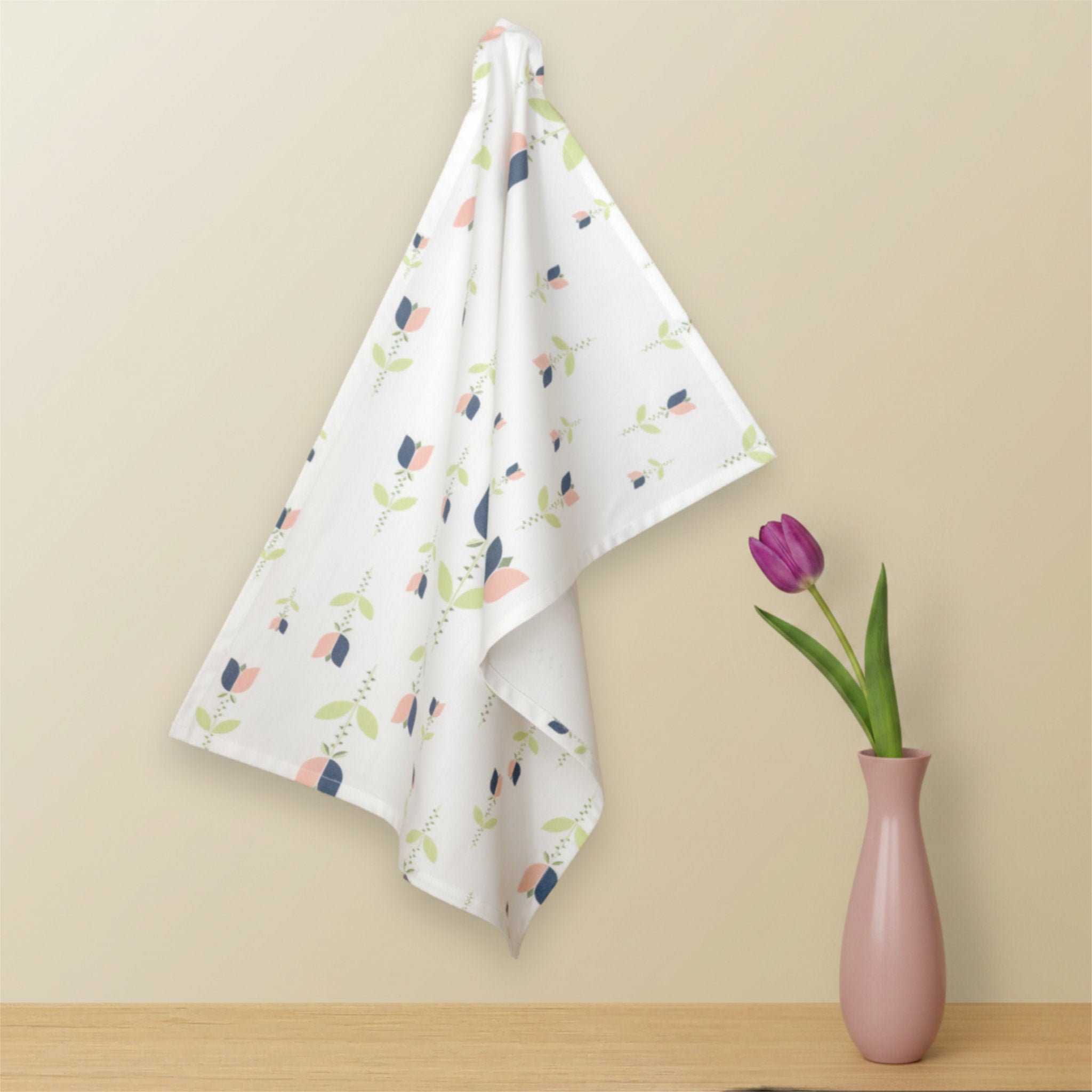 A white kitchen towel with a colorful floral pattern hangs on a wall hook next to a pink vase containing a purple tulip, set against a beige wall and light wood countertop from Hem And Tassel