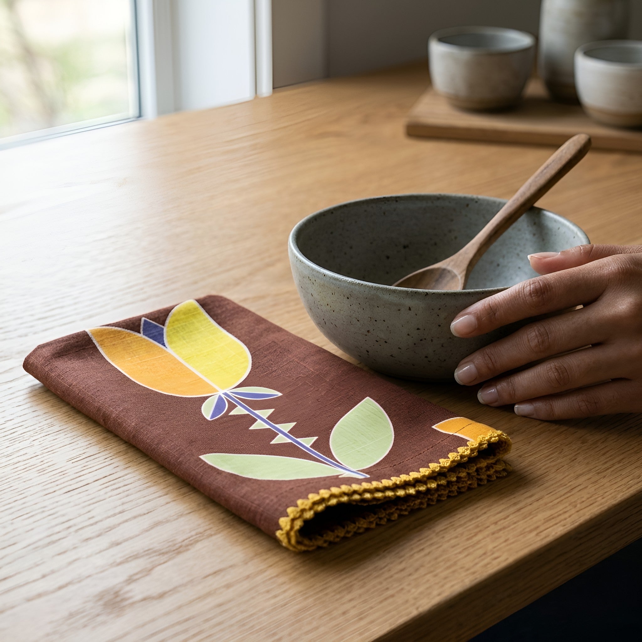 A folded brown fabric napkin with a stylized tulip floral pattern in yellow, green, and blue, featuring a yellow braided edge, is styled on a wooden table next to a speckled gray ceramic bowl and a hand holding a wooden spoon from Hem And Tassel

