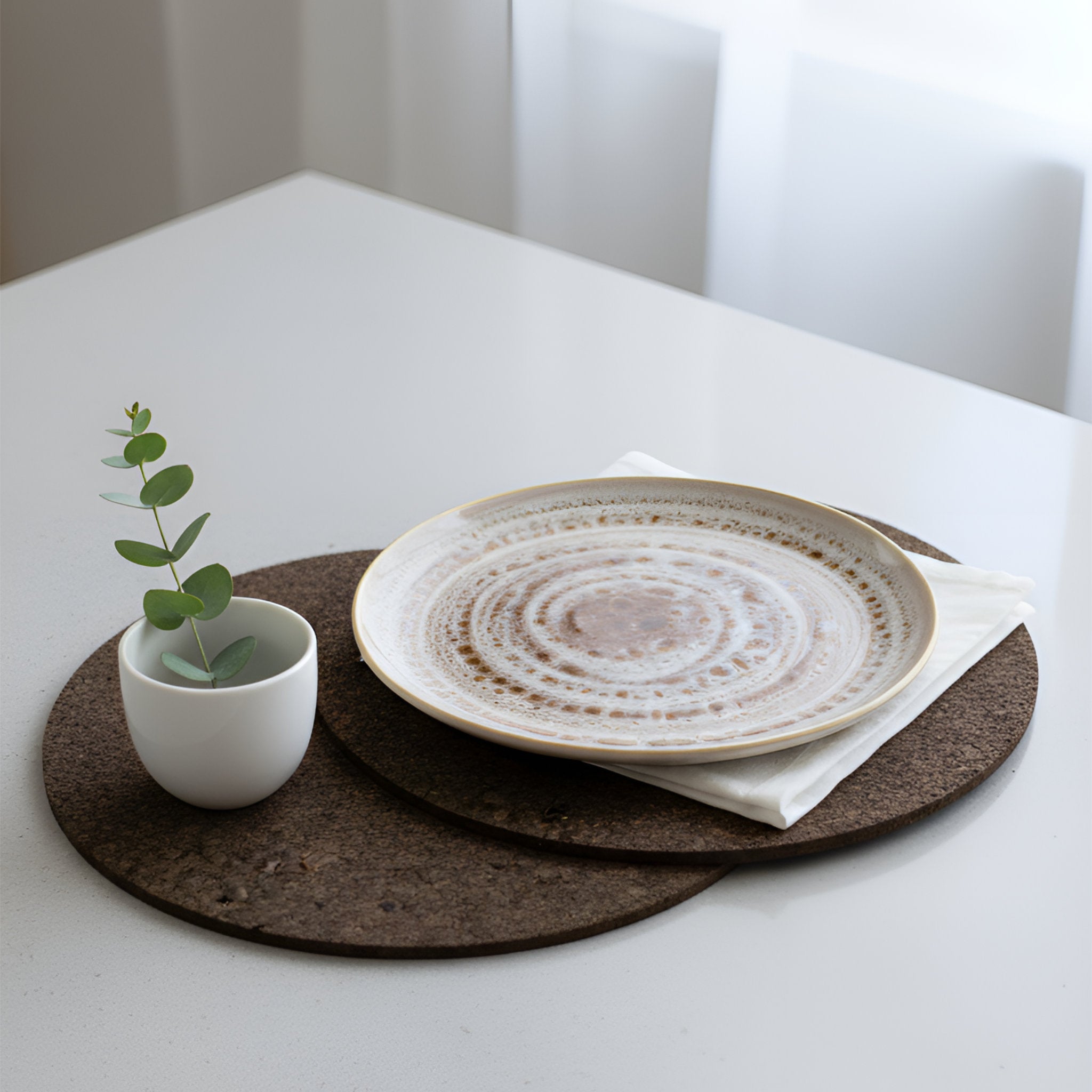 A round ceramic plate with a textured, speckled beige and brown glaze sits on a white napkin atop a dark brown cork placemat on a white table. A small white cup with a green eucalyptus sprig is placed beside it.