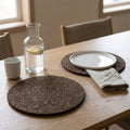 Two round matt black cork placemats on a light wood dining table, styled with a white plate, beige napkin, glass carafe, and white mug in a softly lit room from Hem And Tassel 