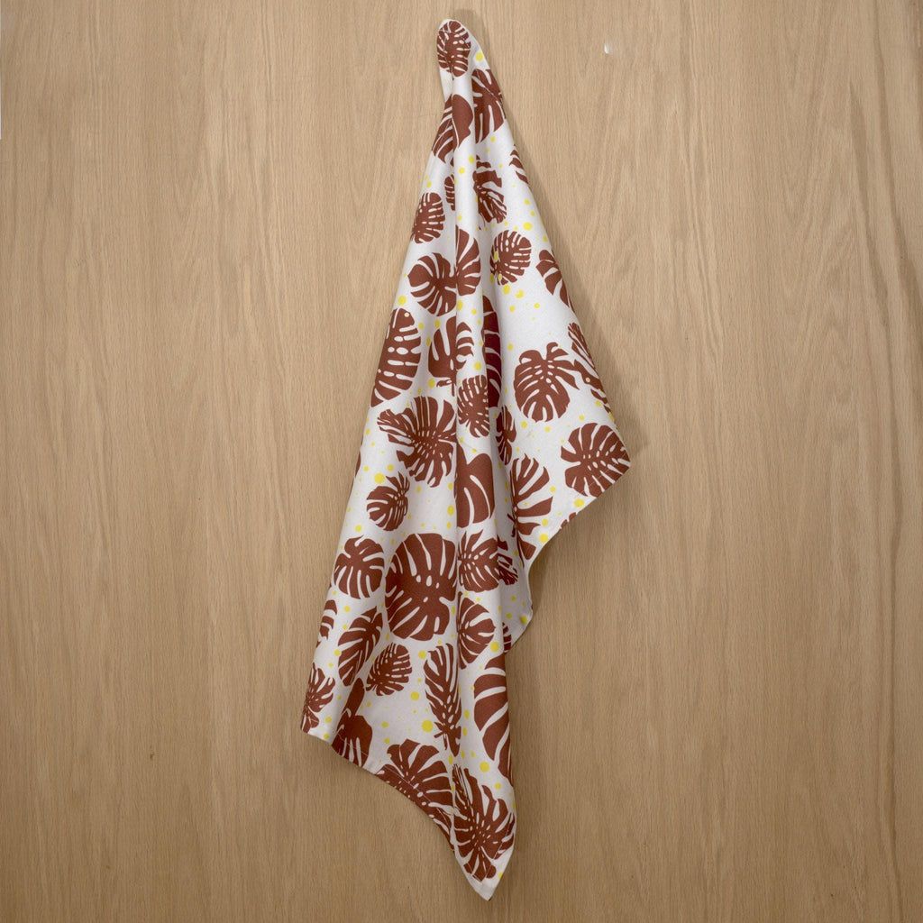 monster ah kitchen towel with brown leaf pattern on a white background hanging against a wooden surface from Hem And Tassel 