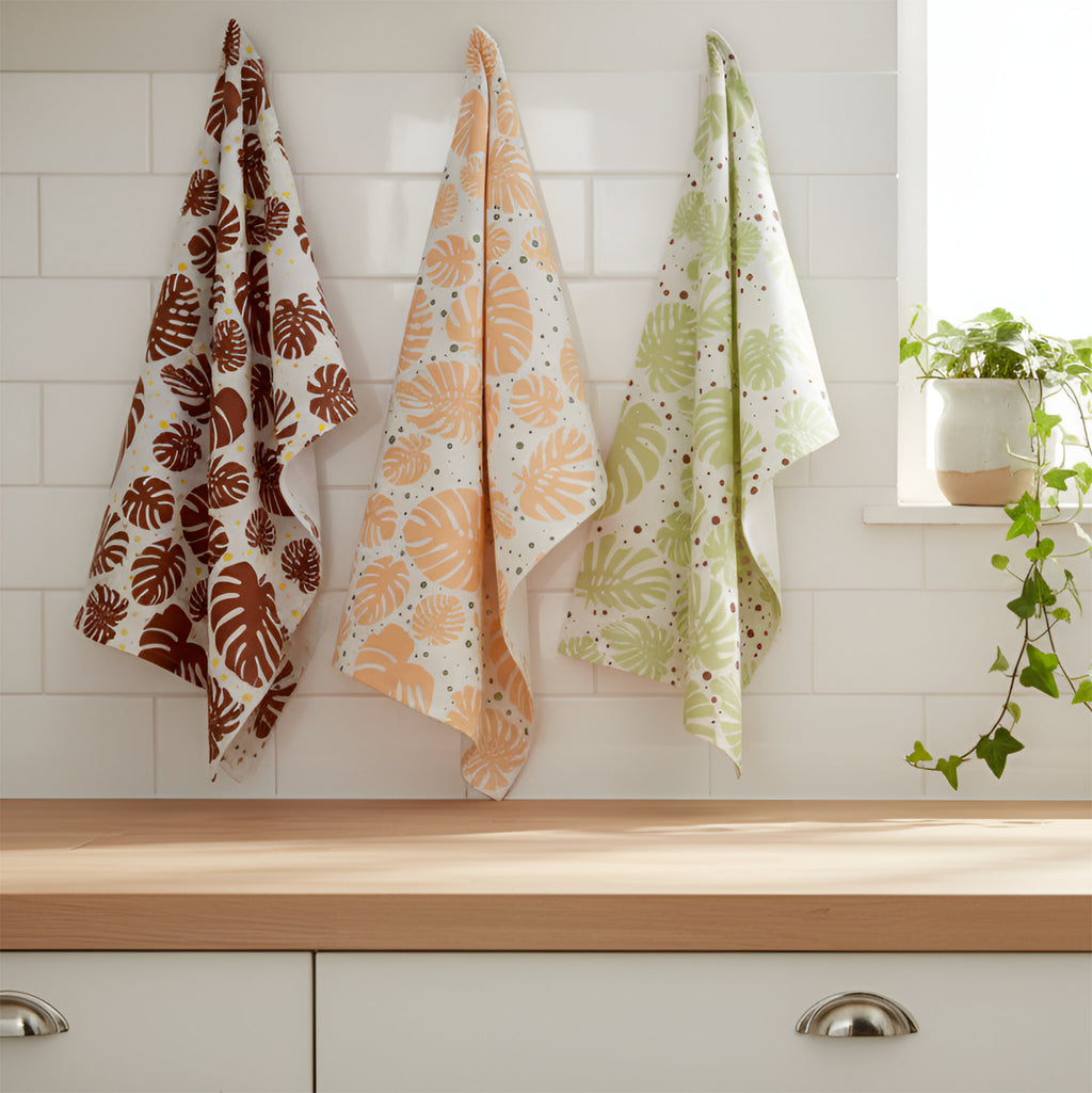 Three monstera leaf patterned kitchen towels hanging above a wooden countertop against a white subway tile wall, with a potted trailing plant on the windowsill. The towels are in brown, peach, and light green on white backgrounds from Hem And Tassel 