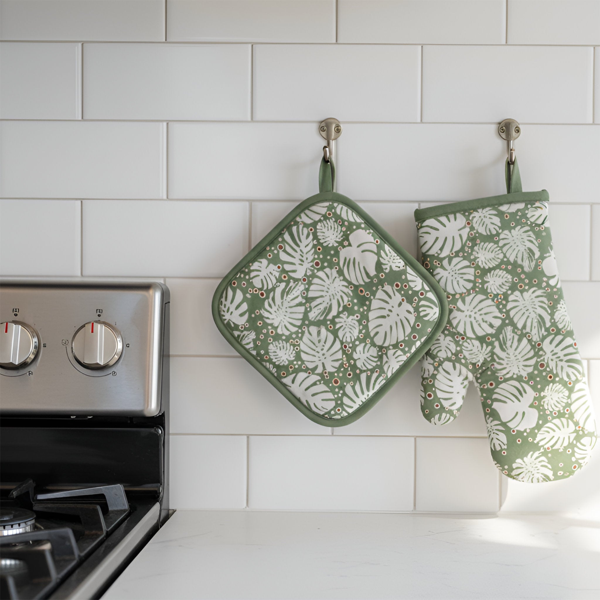 Turf color oven mitt and pot holder of monstera collection hanging on hooks on a white subway tile wall near a cooking range from Hem And Tassel 