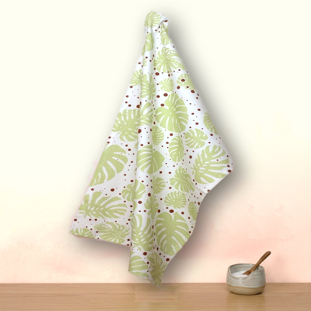 A white kitchen towel with a green monstera leaf pattern and small red polka dots, hanging on a wall above a wooden surface with a small ceramic bowl and wooden spoon from Hem And Tassel 