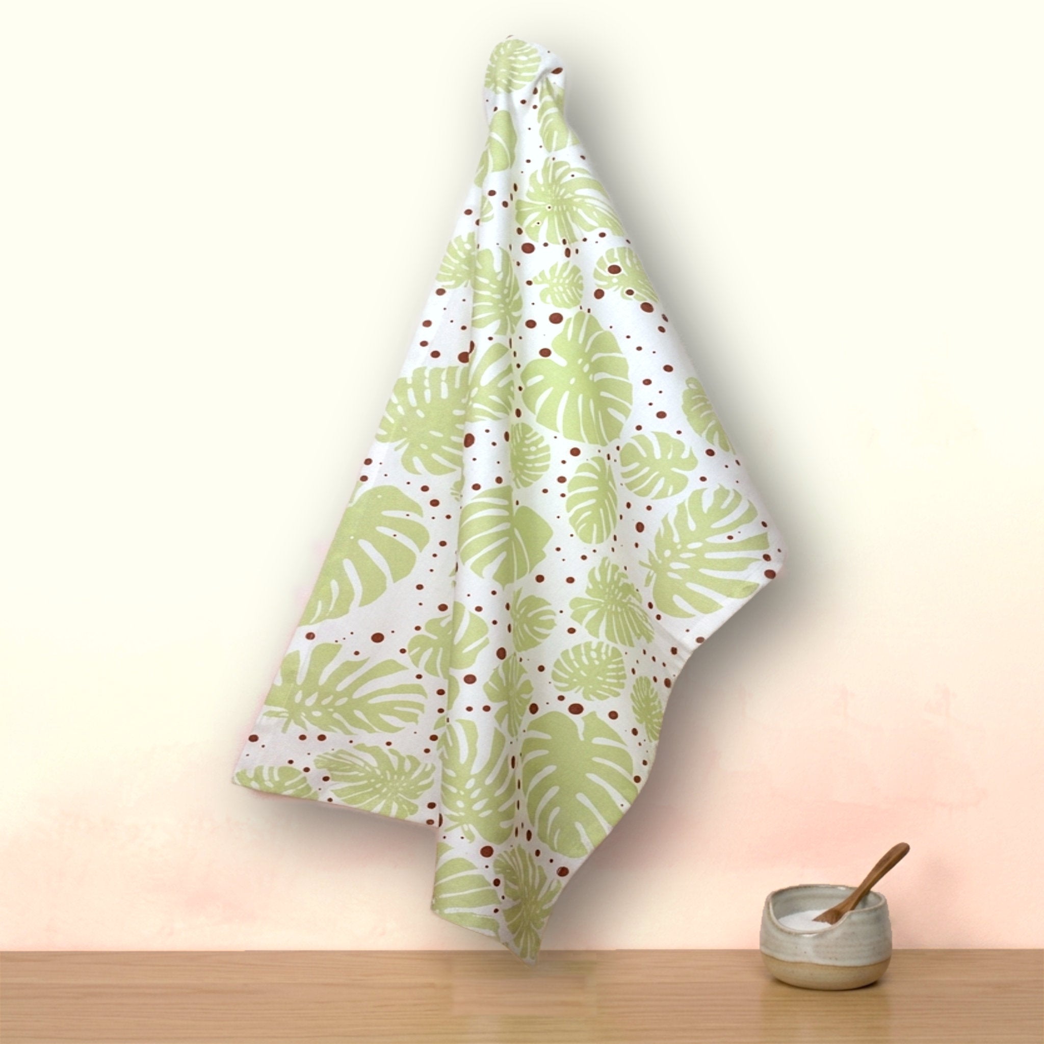 A white kitchen towel with a green monstera leaf pattern and small red polka dots, hanging on a wall above a wooden surface with a small ceramic bowl and wooden spoon from Hem And Tassel 