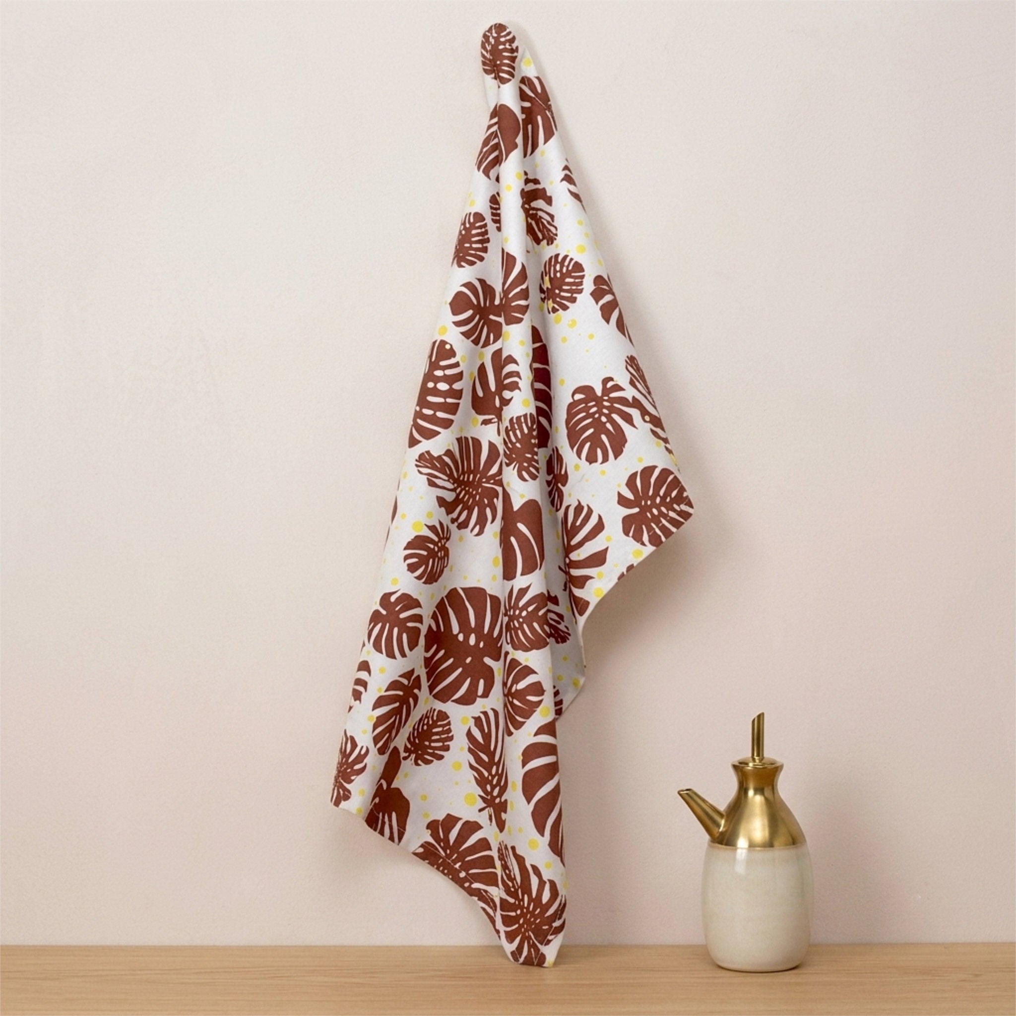 A white kitchen towel with a brown monstera leaf pattern hangs from a wall hook next to a small ceramic and brass pitcher on a light wood surface from Hem And Tassel 