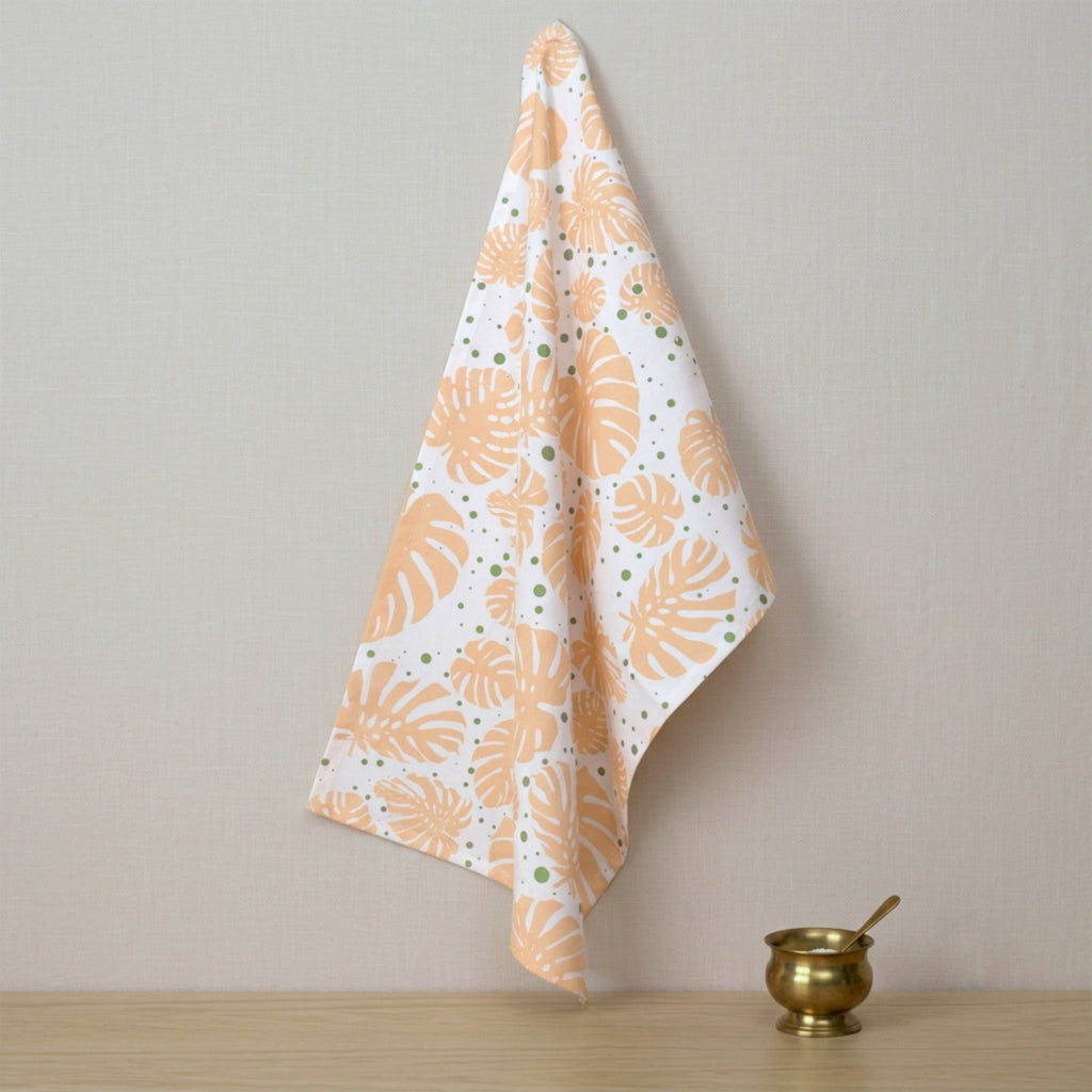 A white kitchen towel with a pattern of orange monstera leaves and small green dots, hanging on a wall next to a small brass bowl on a wooden surface from Hem And Tassel 