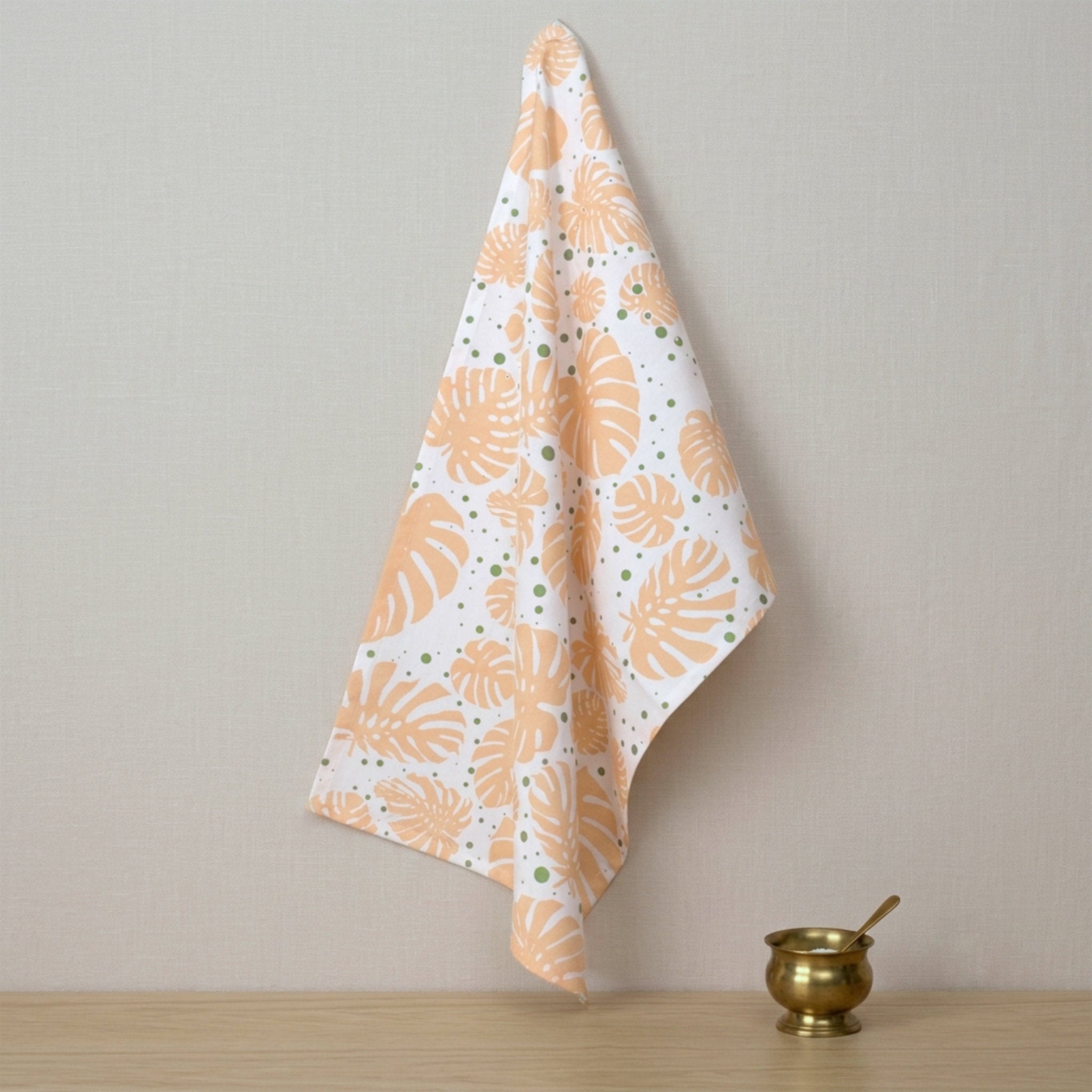A white kitchen towel with a pattern of orange monstera leaves and small green dots, hanging on a wall next to a small brass bowl on a wooden surface from Hem And Tassel 
