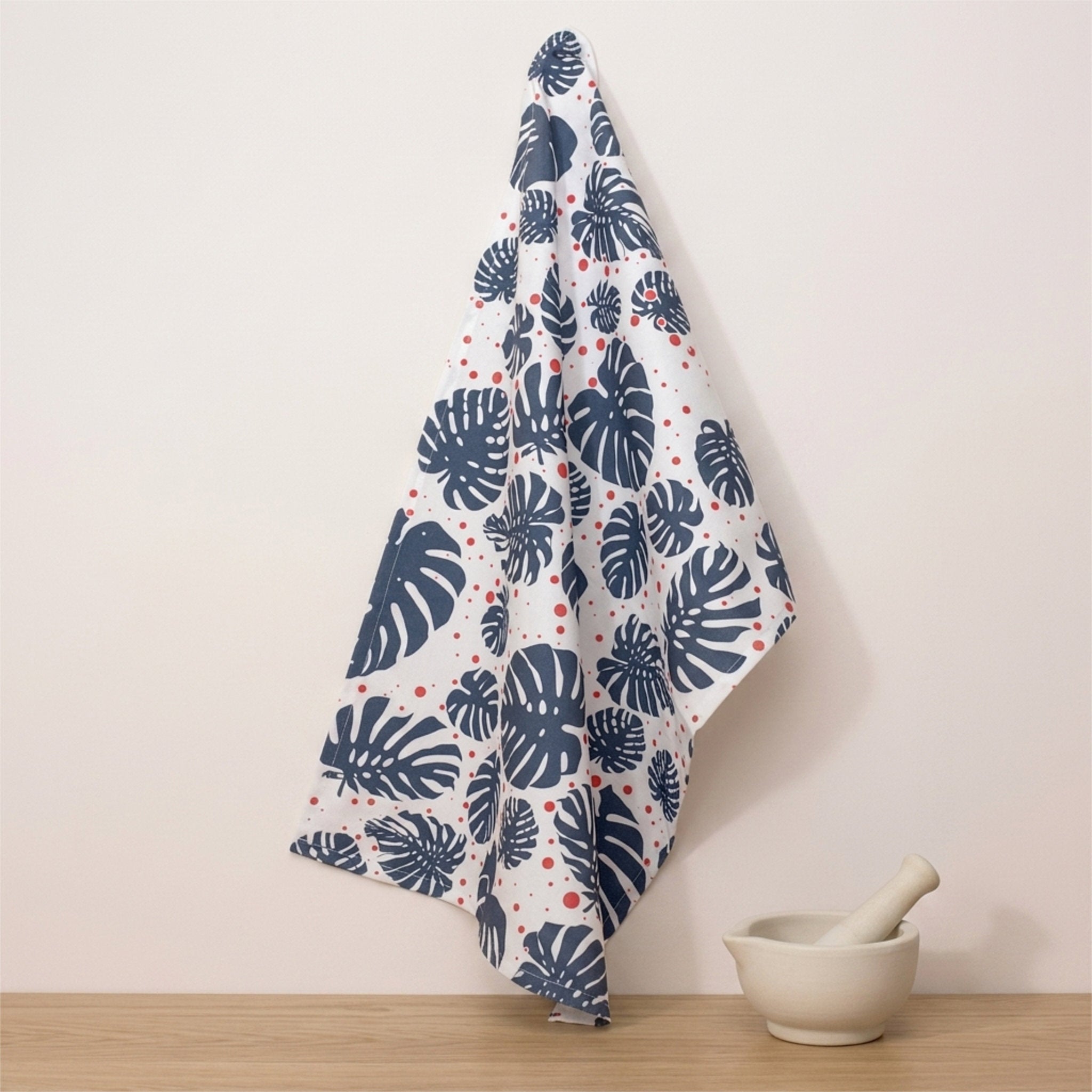 A white kitchen towel with a navy blue monstera leaf pattern and small red polka dots, hanging on a wall hook above a wooden surface next to a ceramic mortar and pestle from Hem And Tassel 