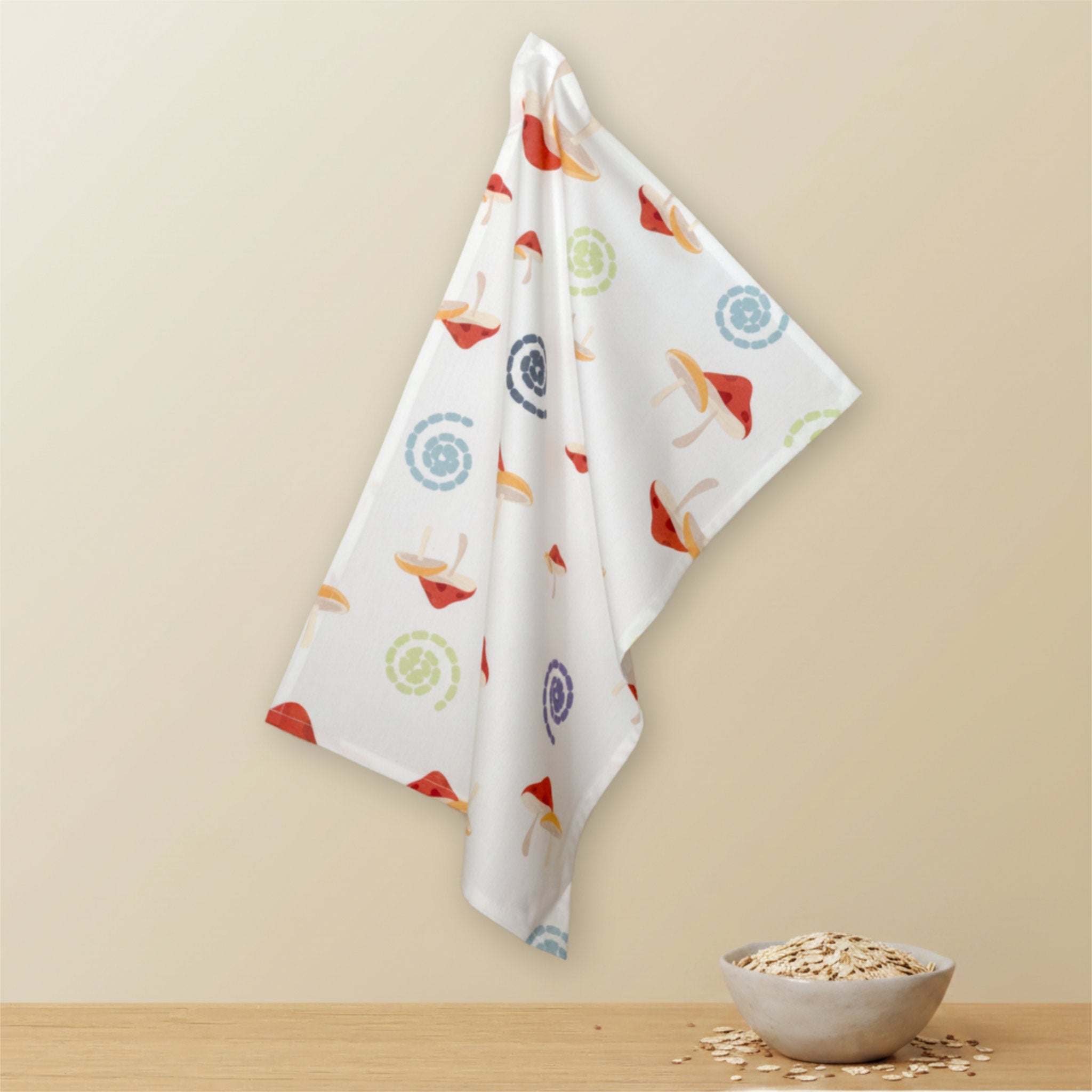 A white kitchen towel with a colorful mushroom and swirl pattern hangs against a beige wall above a wooden surface with a small bowl of oats from Hem And Tassel