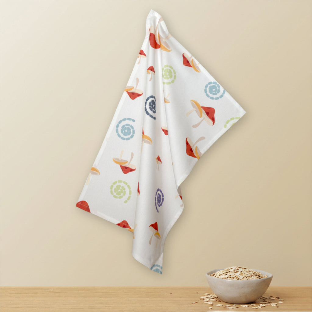 A white kitchen towel with a colorful mushroom and swirl pattern hangs against a beige wall above a wooden surface with a small bowl of oats from Hem And Tassel