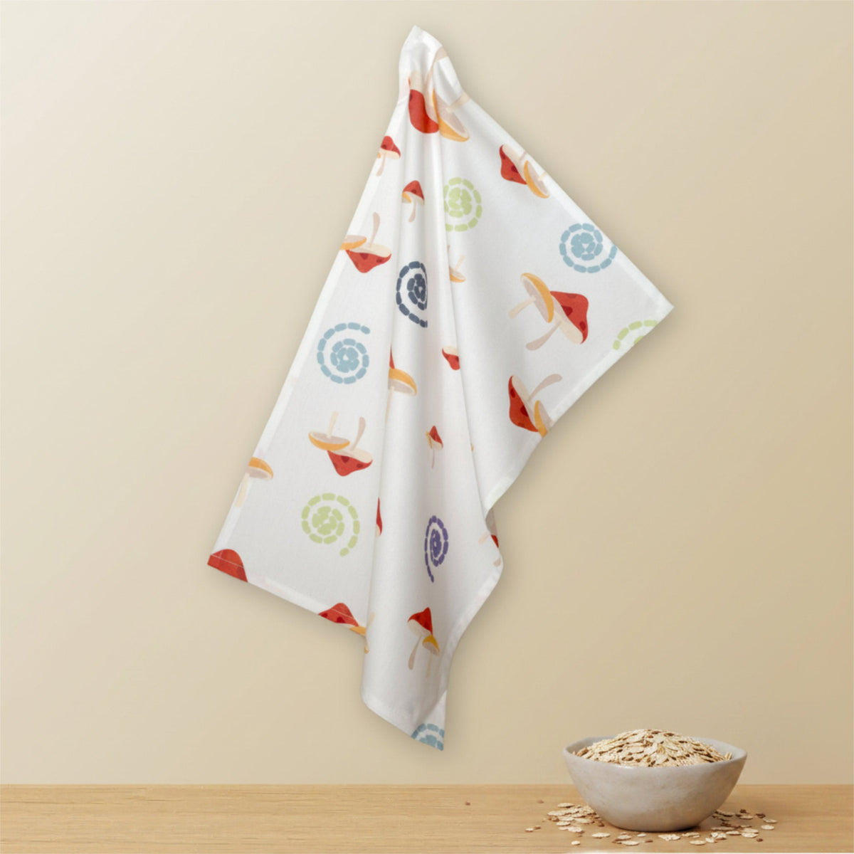 A white kitchen towel with a colorful mushroom and swirl pattern hangs against a beige wall above a wooden surface with a small bowl of oats from Hem And Tassel
