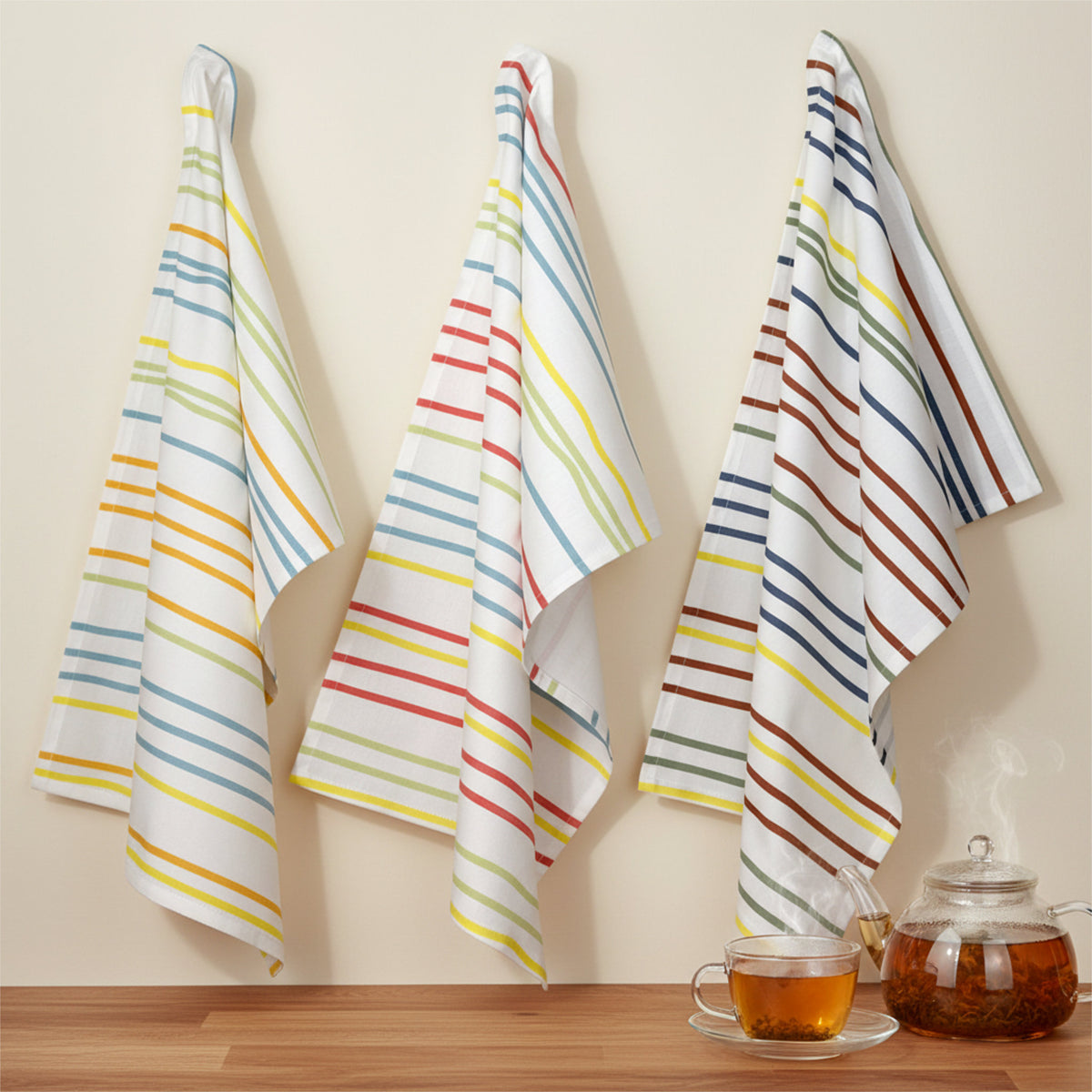 narrow-width stripes in Spring, summer and Fall colors on 3 cotton kitchen towels hanging on a beige wall with a pot of herbal tea, cup and saucer on a wood counter below from Hem And Tassel 