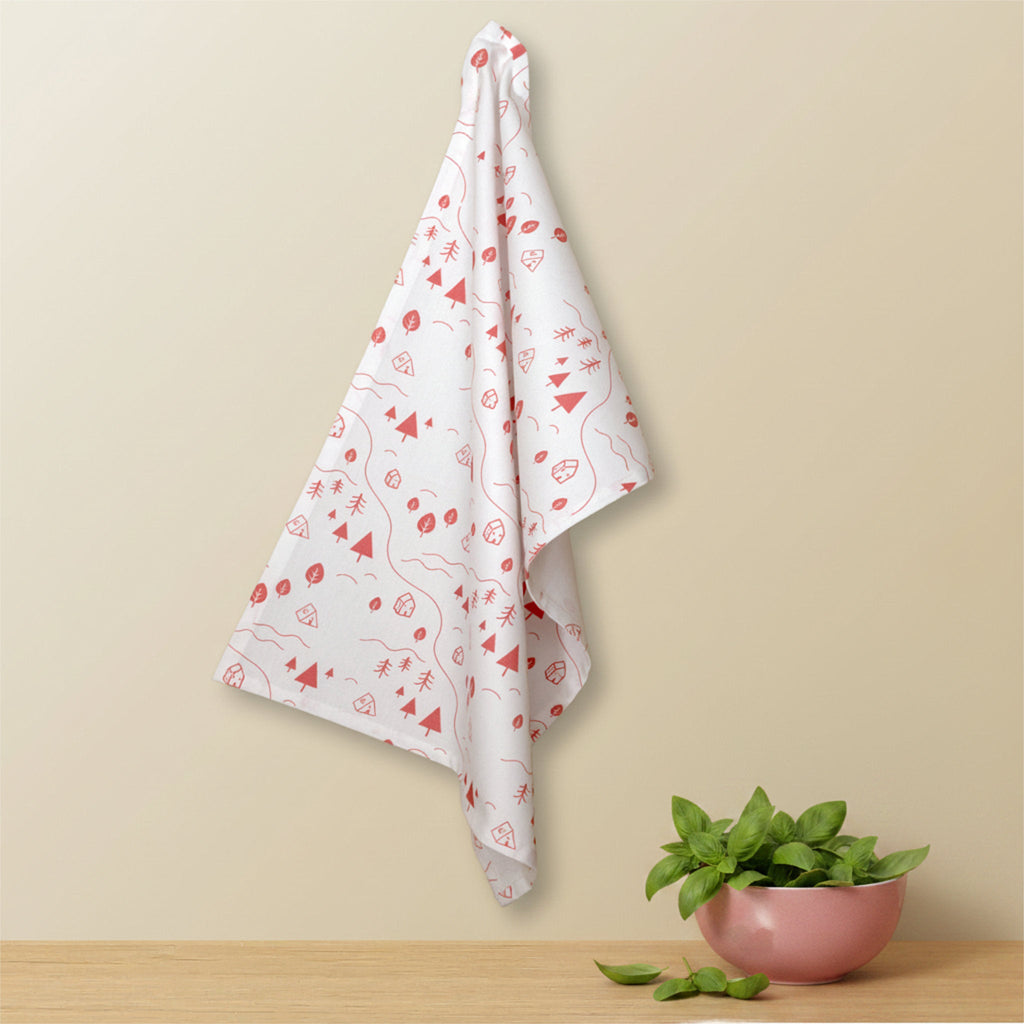 A white kitchen towel with a pink minimalist pattern of trees, tents, leaves, and abstract shapes, hanging on a beige wall above a wooden table with a small pink bowl of basil from Hem And Tassel