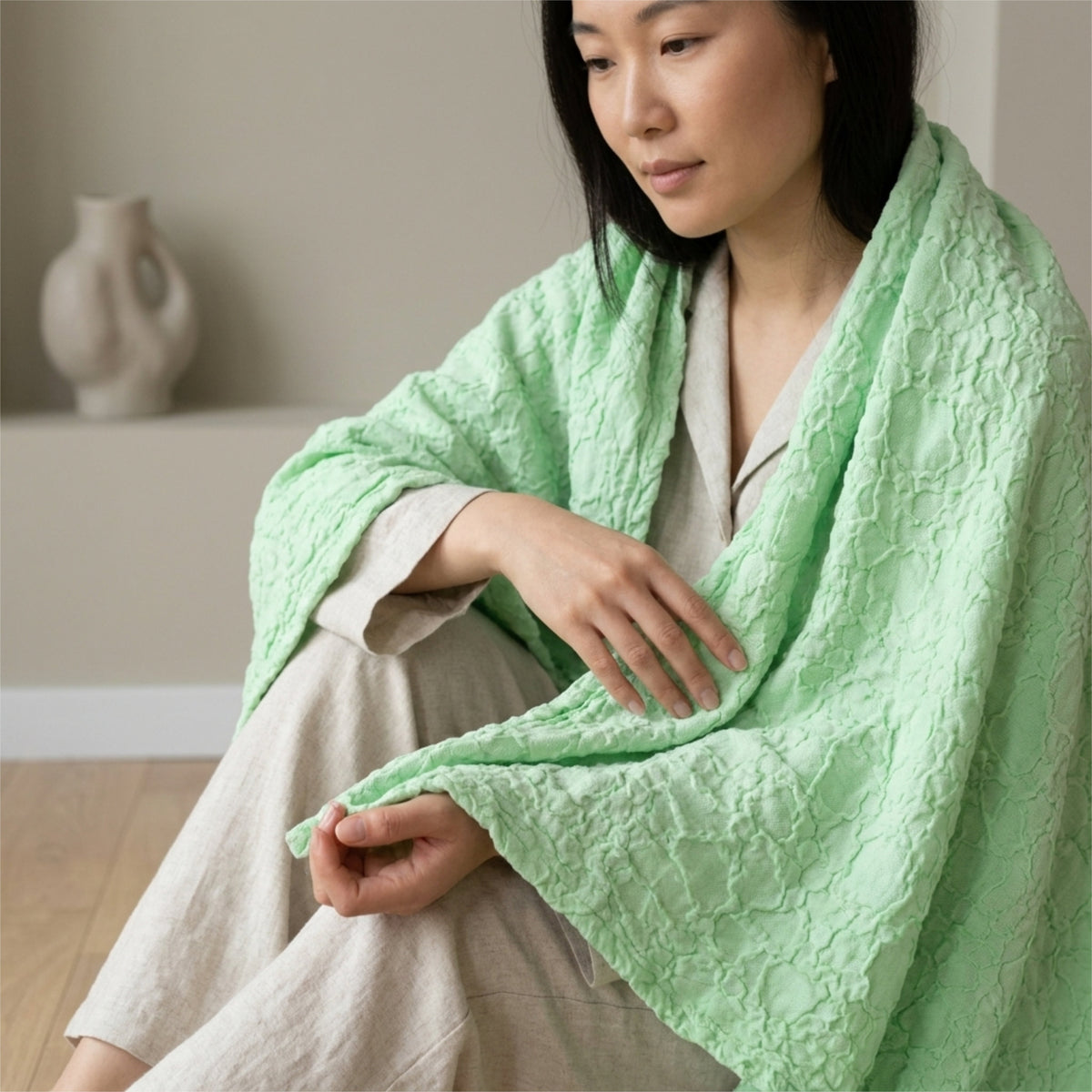 A woman seated indoors wearing a box waffle soft mint colored woven throw blanket hemmed on four sides. The background is softly blurred with a neutral wall and a small vase from Hem And Tassel 