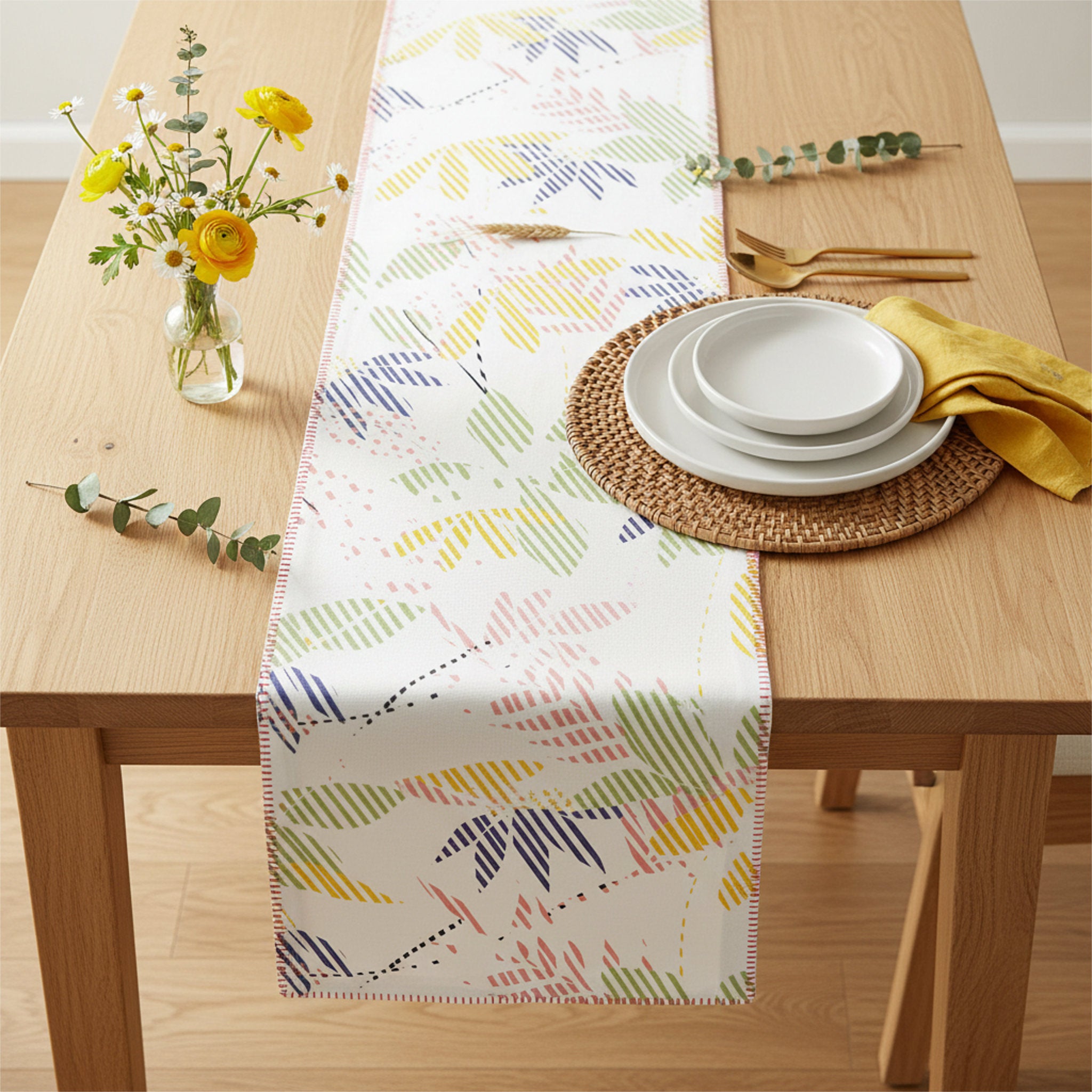 A white table runner with a colorful abstract leaf pattern is styled on a light wood dining table. The table setting includes a woven placemat, white plates, a yellow napkin, gold-toned cutlery, and a small glass vase with yellow flowers and greenery from Hem And Tassel 