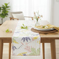 A white table runner with a colorful palm leaf pattern is styled on a light wood dining table, surrounded by plates, a yellow napkin, and small flowers in a vase from Hem And Tassel 