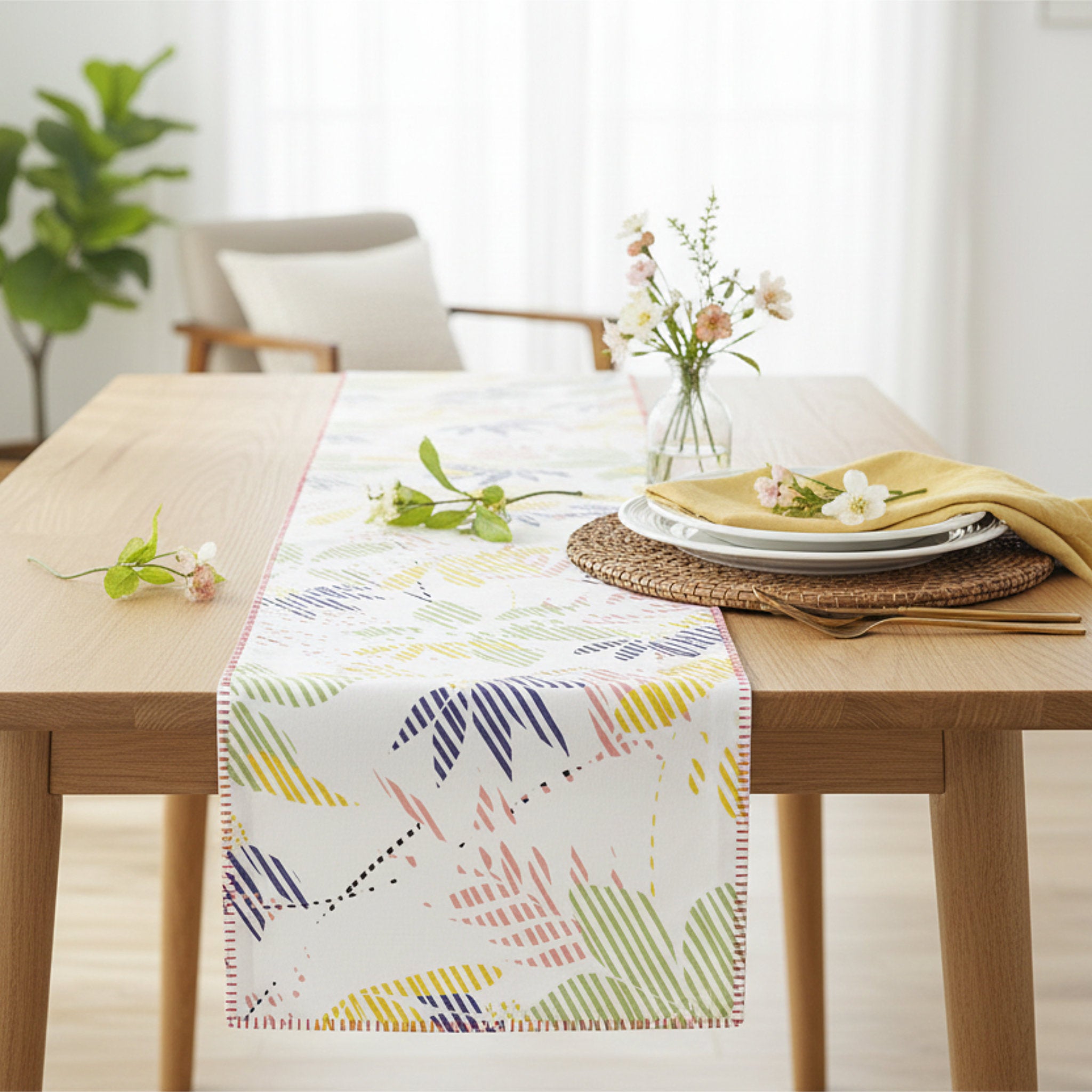 A white table runner with a colorful palm leaf pattern is styled on a light wood dining table, surrounded by plates, a yellow napkin, and small flowers in a vase from Hem And Tassel 