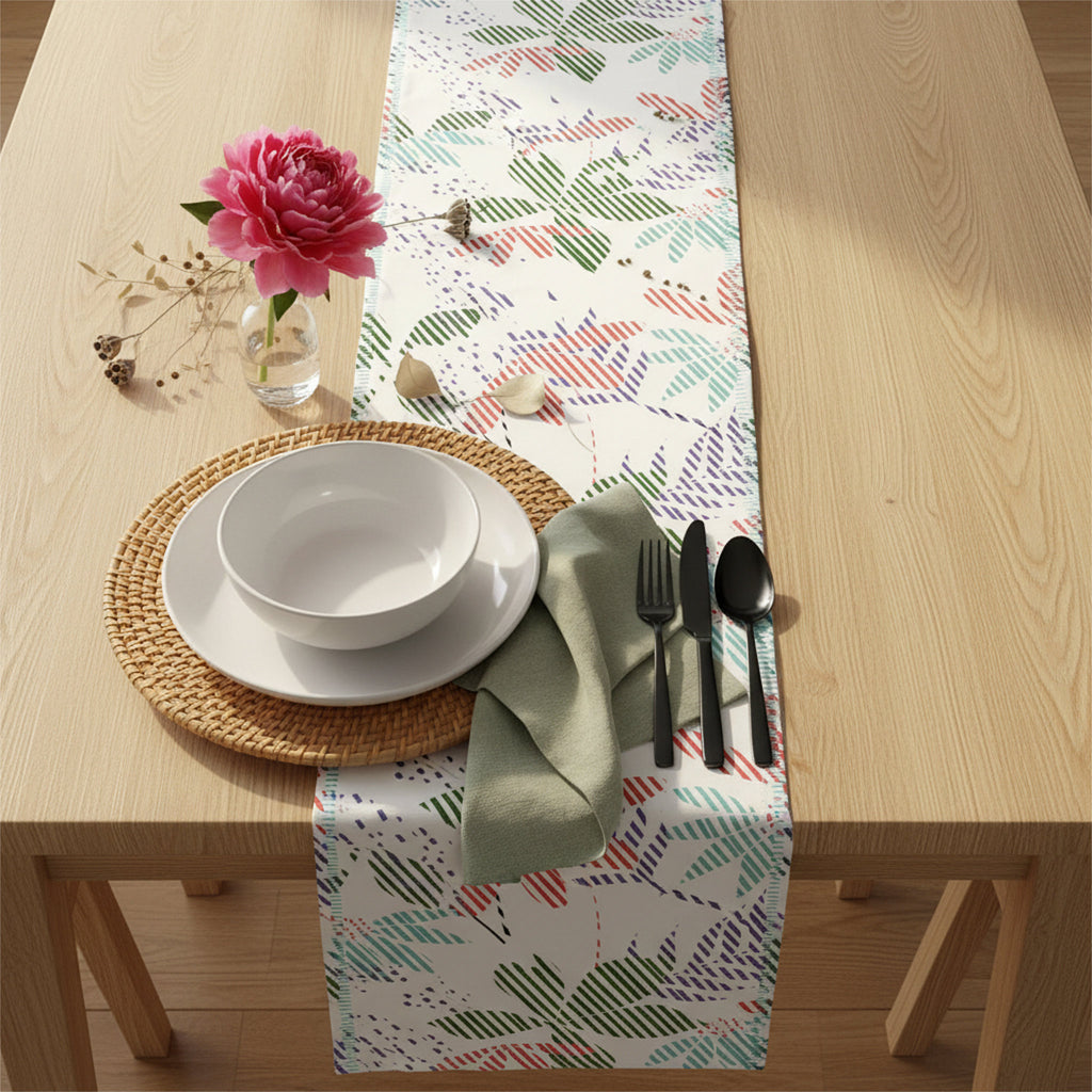 A rectangular table runner with a colorful botanical leaf pattern in green, red, and blue on a white background, styled on a light wood dining table with a place setting, a pink flower, and cutlery from Hem And Tassel 