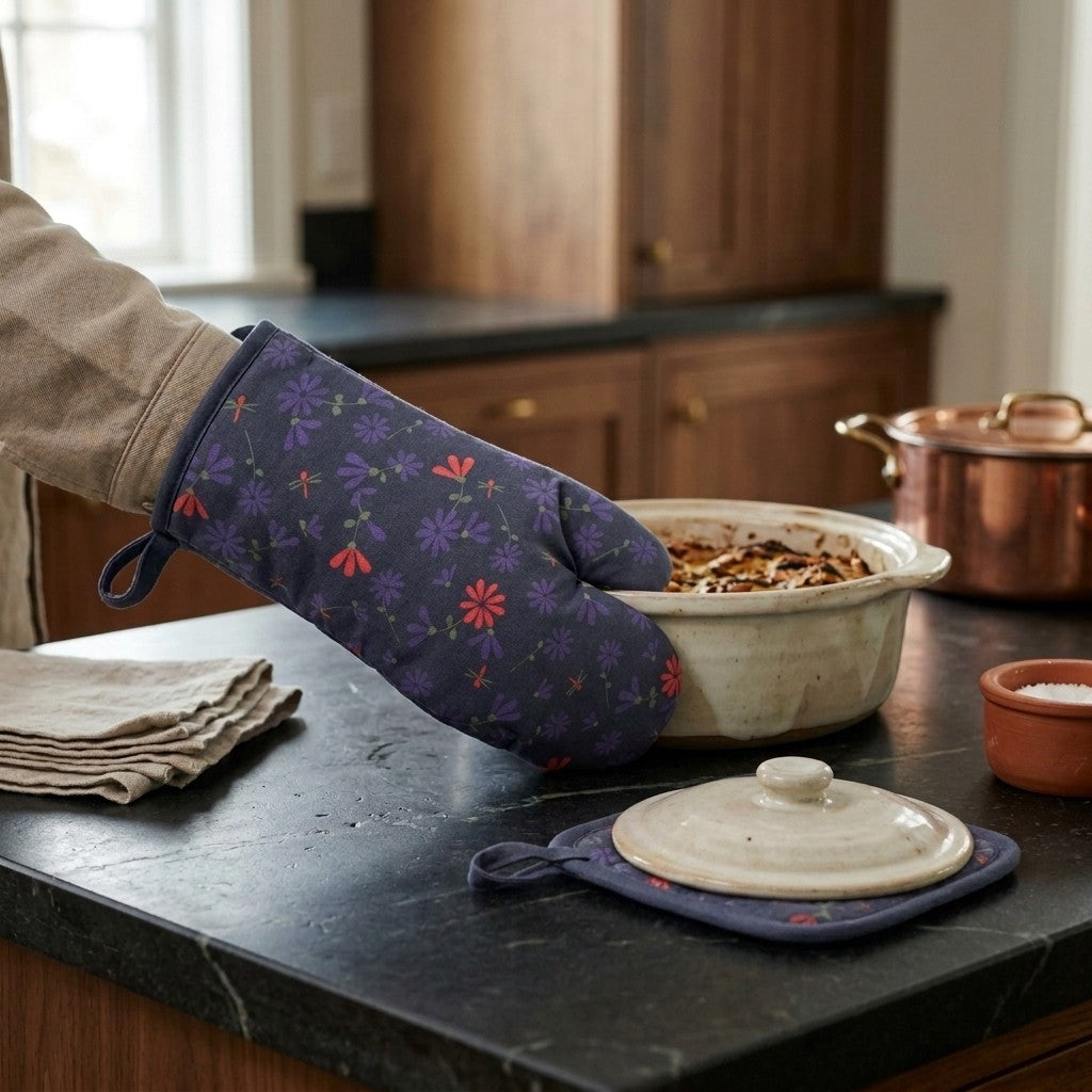 a hand holds a hot casserole with food in it using a dark oil colored oven mitt with colorful floral print of the shy dragonfly pattern while the hot lid sits on the matching pot holder using it as a trivet, on a dark kitchen counter from Hem And Tassel