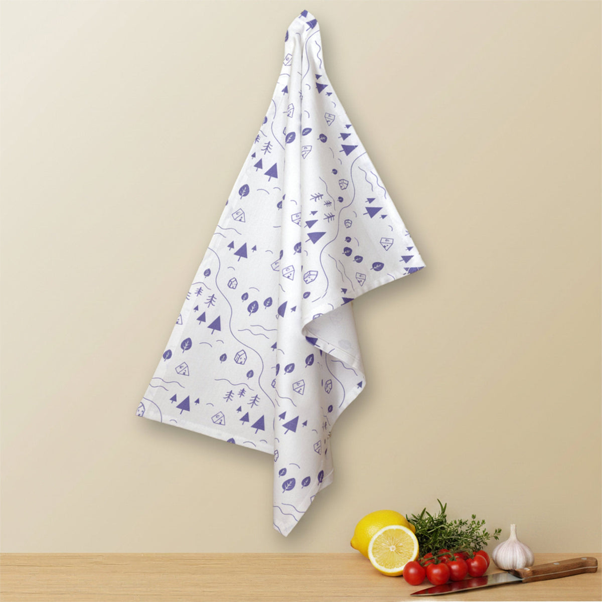 A cotton kitchen towel with a colorful tiny countryside icons all over on a white background, hanging against a beige wall above a wooden surface with a cut lemon, herbs, garlic, cherry tomatoes and a knicfe from Hem And Tassel