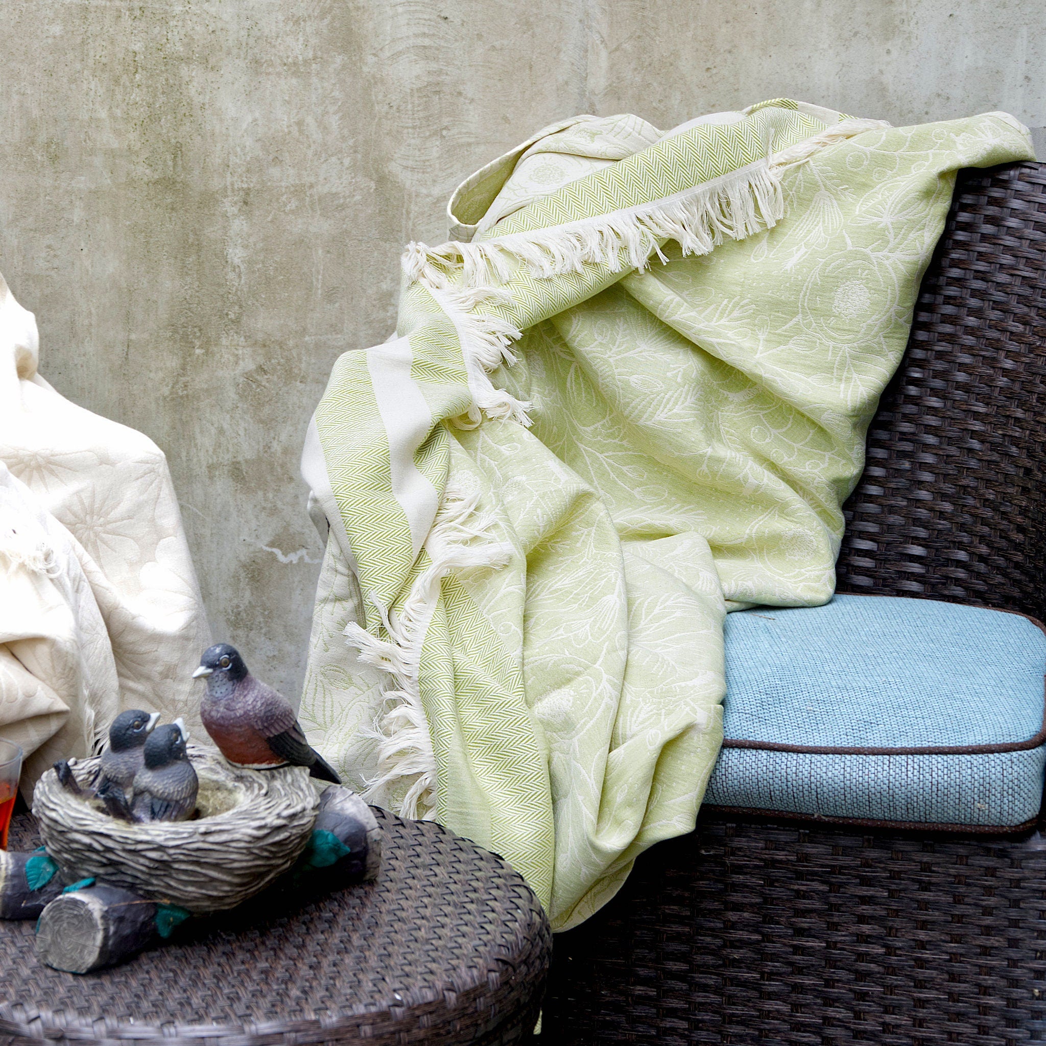 Light green throw blanket draped over a wicker chair with a small bird nest and birds on a table. from hem and tassel