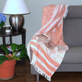 Coral and white patterned throw blanket draped over a gray sofa with a side table and lamp in the background from hem and tasel