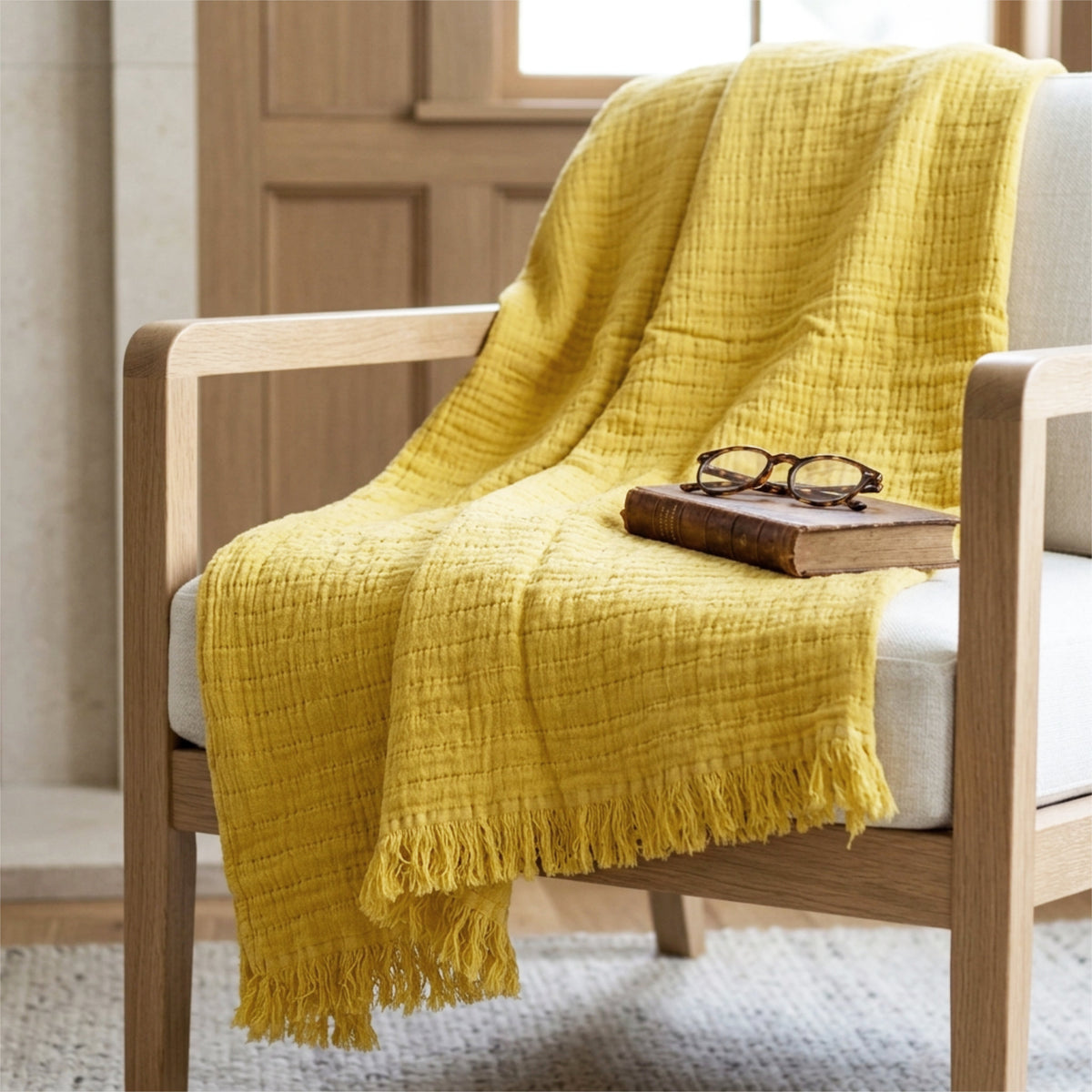 A mustard yellow seersucker throw blanket with tassels is draped over a light wood armchair with a white seat cushion. A vintage book and eyeglasses rest on the chair, set in a cozy room with a wooden door and window in the background from Hem And Tassel 