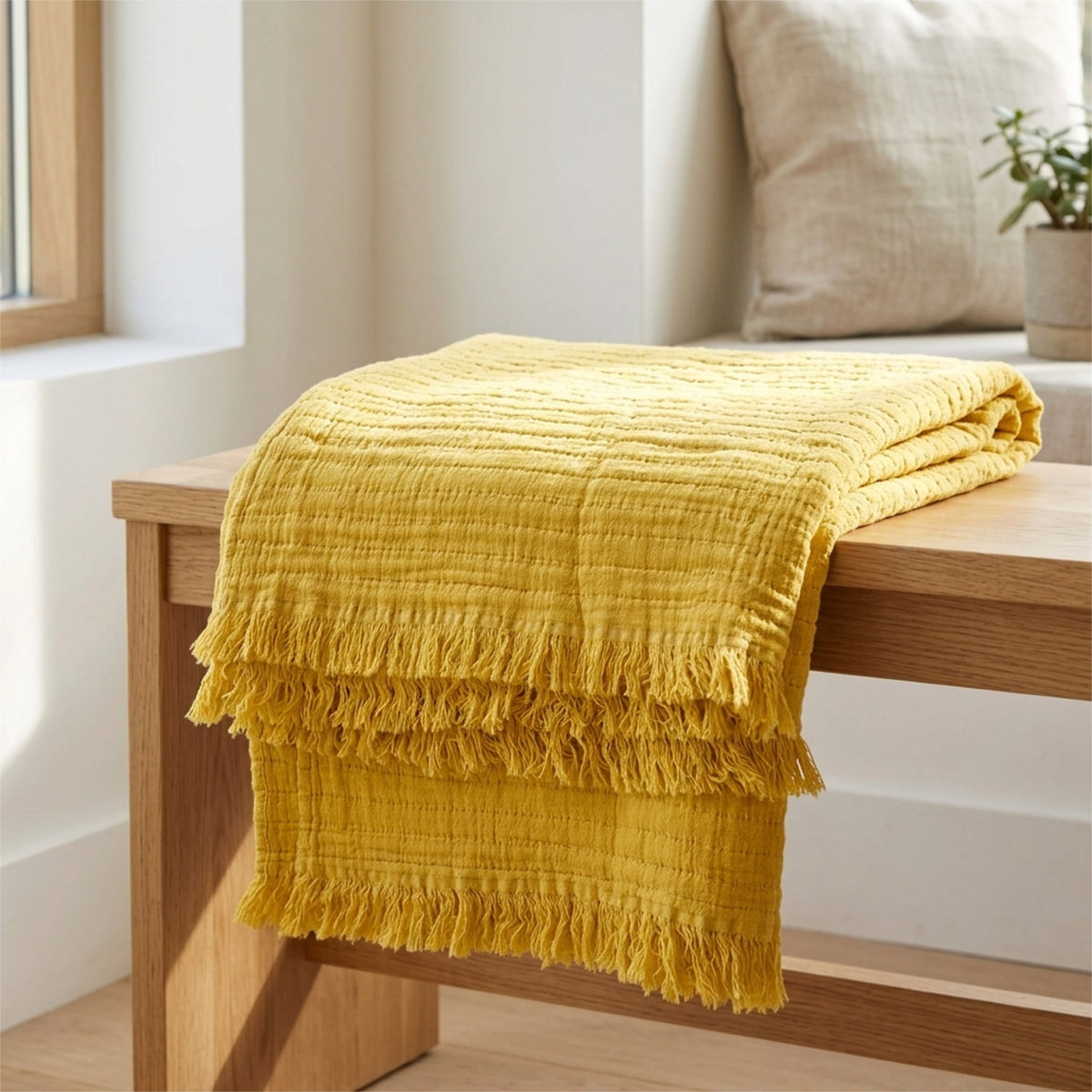 A folded mustard yellow seersucker throw blanket with tasseled trim draped over a light wood bench in a bright, modern room from Hem And Tassel 