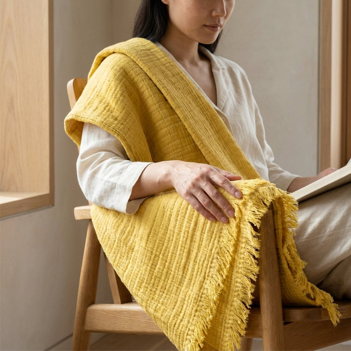 A woman seated on a wooden chair indoors, wrapped in a mustard yellow seersucker throw with tassels. She wears a light cream blouse and beige pants, and the room has warm neutral walls and wood trim from Hem And Tassel 