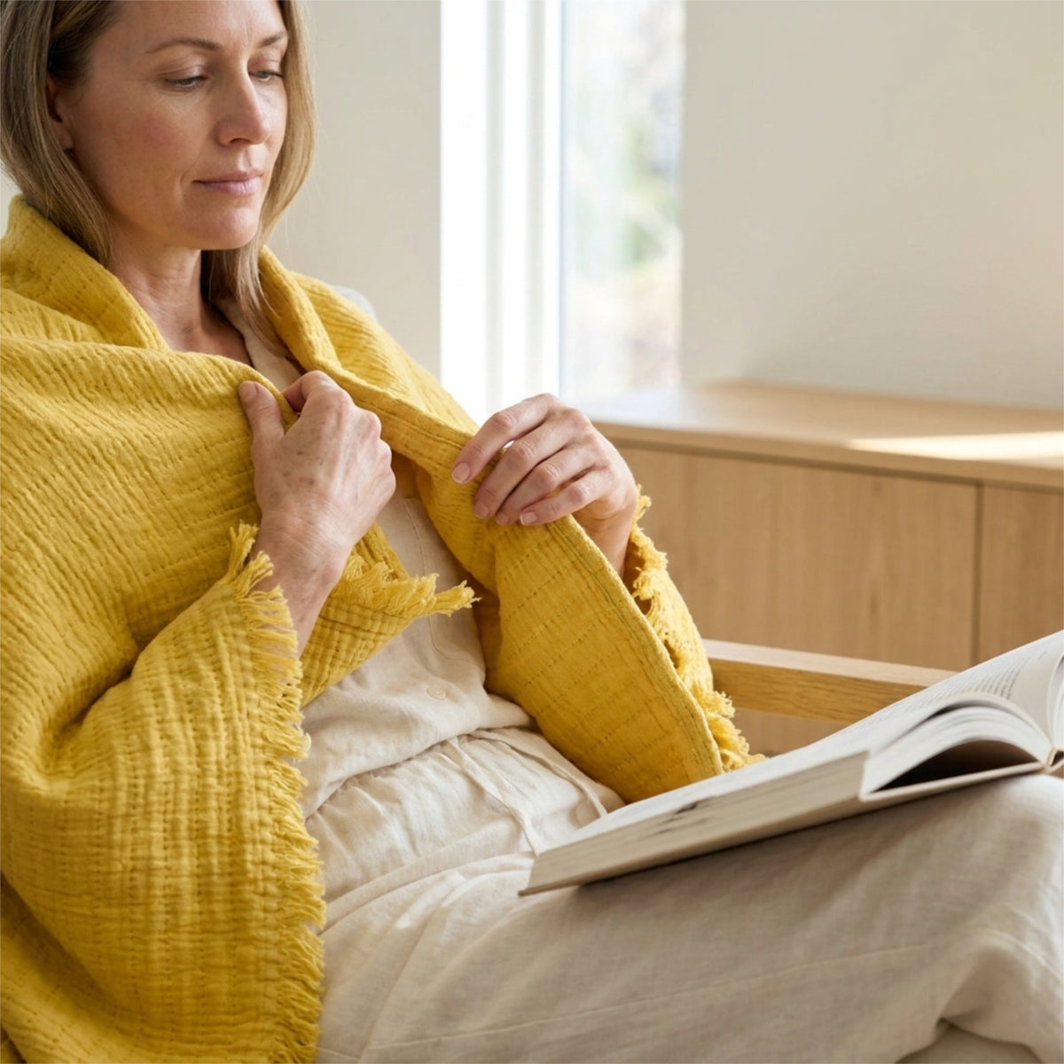 A woman wrapped in a ochre yellow seersucker throw sits indoors near a window, holding an open book from Hem And Tassel 