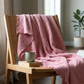 A dusty pink seersucker throw with tasseled edges draped over a light wood chair in a softly lit room, with a ceramic mug resting on the chair seat from Hem And Tassel 