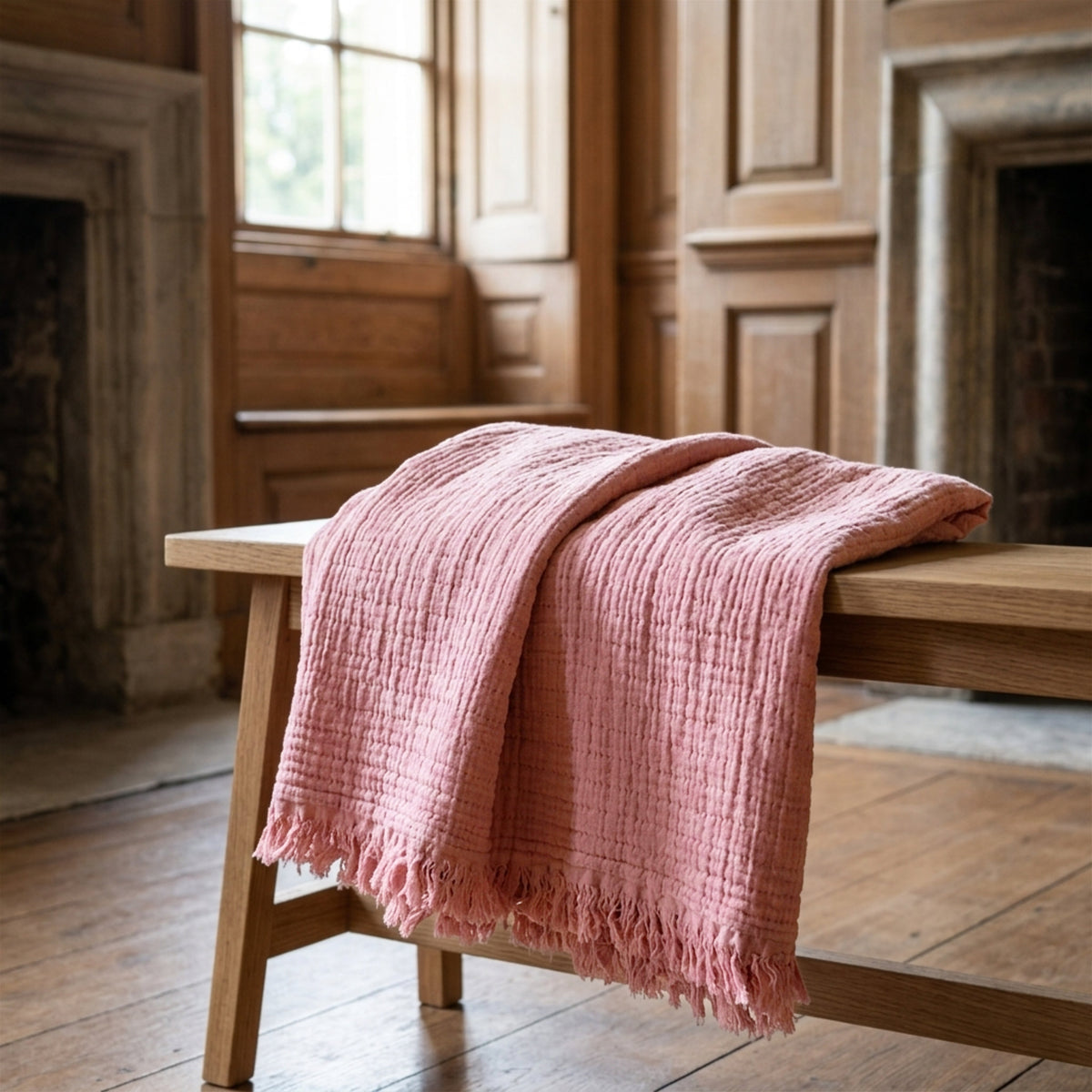 A dusty pink seersucker throw blanket with tasseled edges draped over a wooden bench in a warm, wood-paneled room from Hem And Tassel 