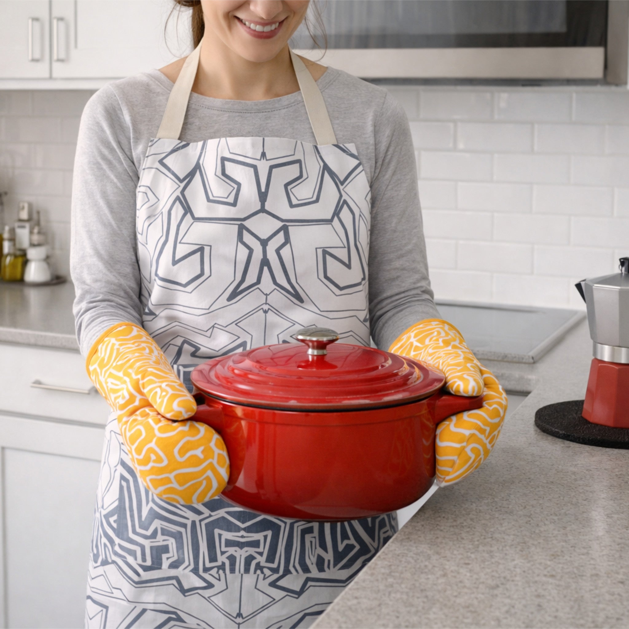 Person wearing a patterned apron and yellow oven mitts holding a red pot in a kitchen.