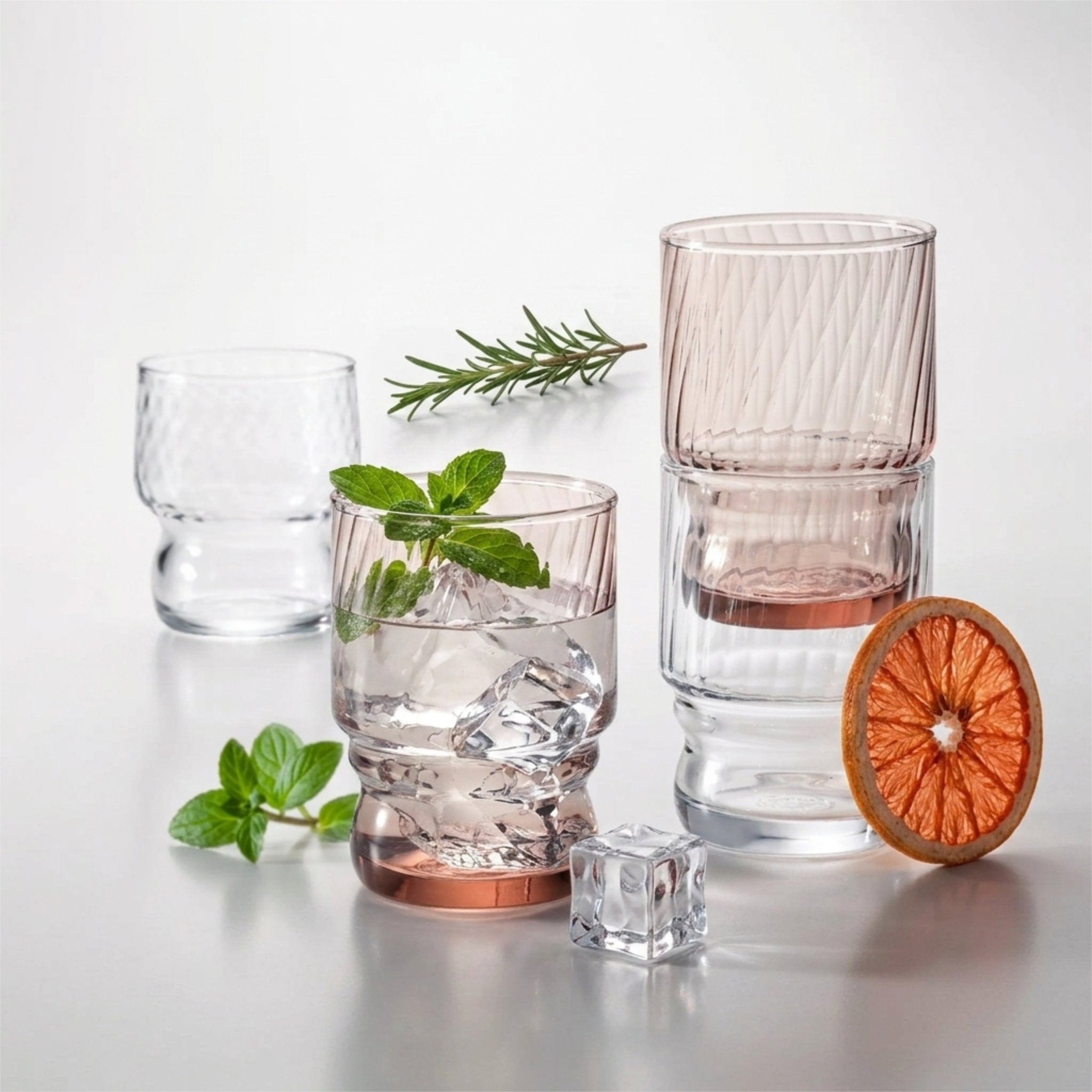 Four textured clear stackable drinking glasses with a subtle pink tint, styled with mint leaves, a dried orange slice, and ice cubes on a reflective light gray surface from Hem And Tassel 