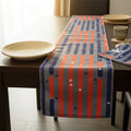 A striped table runner with a patriotic red, white, and blue design is styled on a dark wooden dining table alongside ceramic plates and a matching napkin from Hem And Tassel 