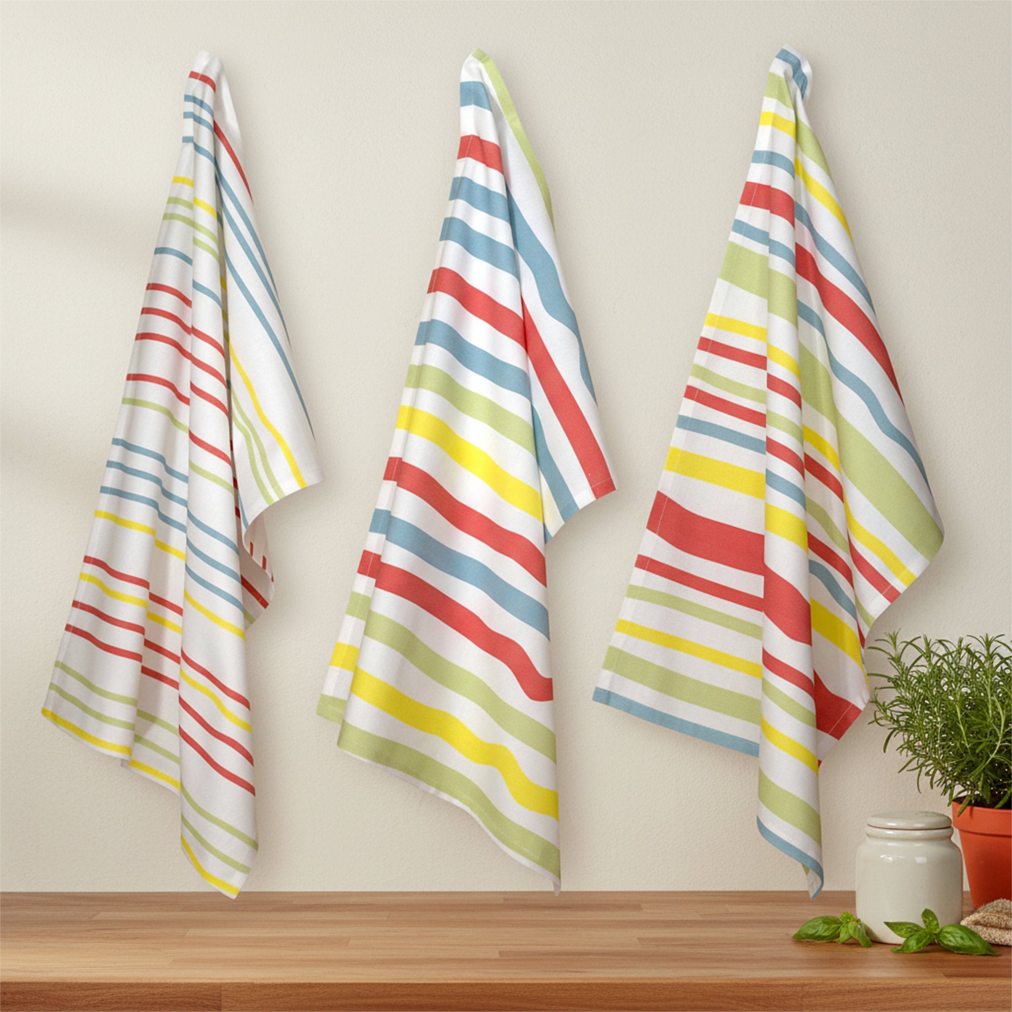 Summer colors stripes in narrow, medium and broad widths on 3 cotton kitchen towels hanging on a beige wall with a pot of rosemary and some basil leaves on a wood counter below from Hem And Tassel 