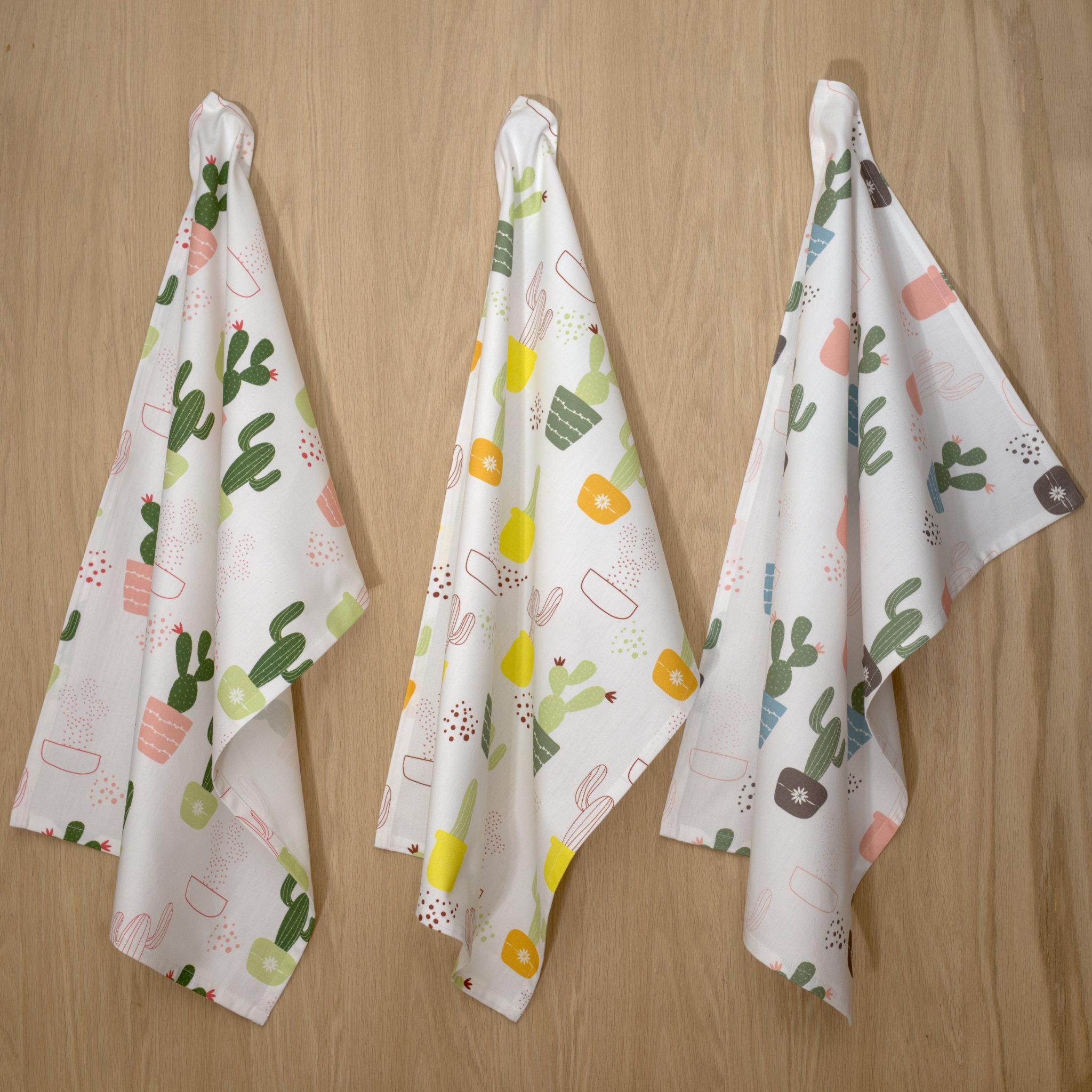 Three decorative thorny issue kitchen towels with cactus patterns hanging on a wooden wall from Hem And Tassel 
