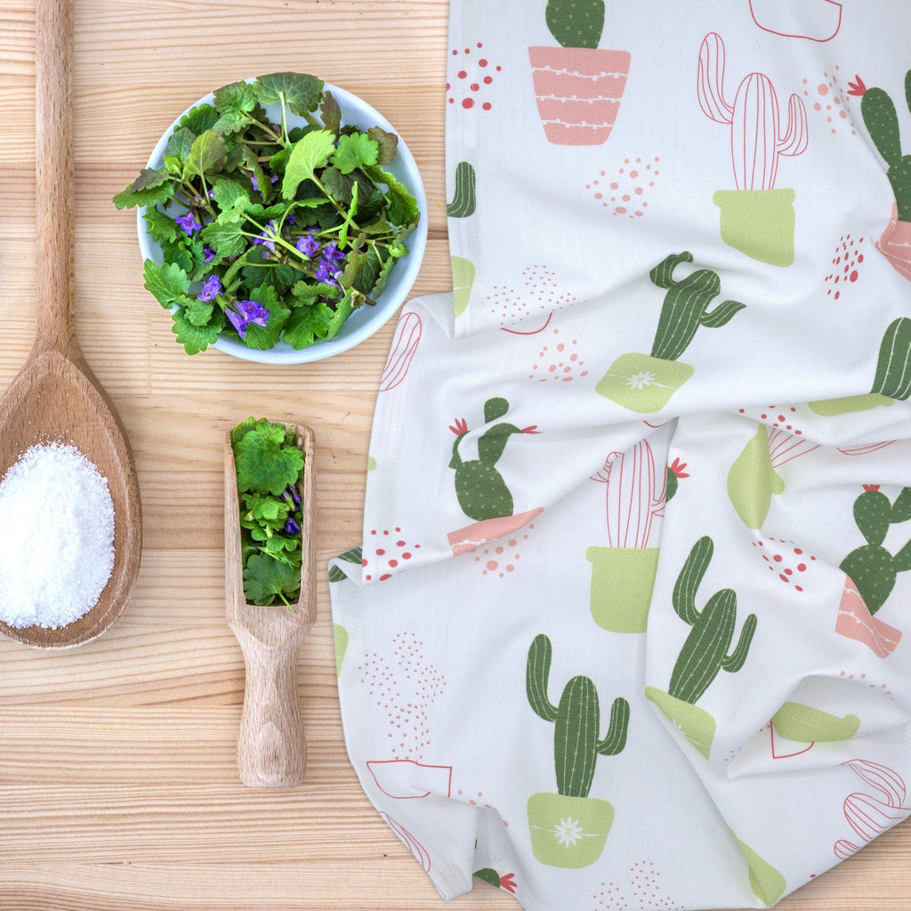 Cactus-patterned thorny issue kitchen towel on a wooden surface with a bowl of greens and a wooden spoon from Hem And Tassel 
