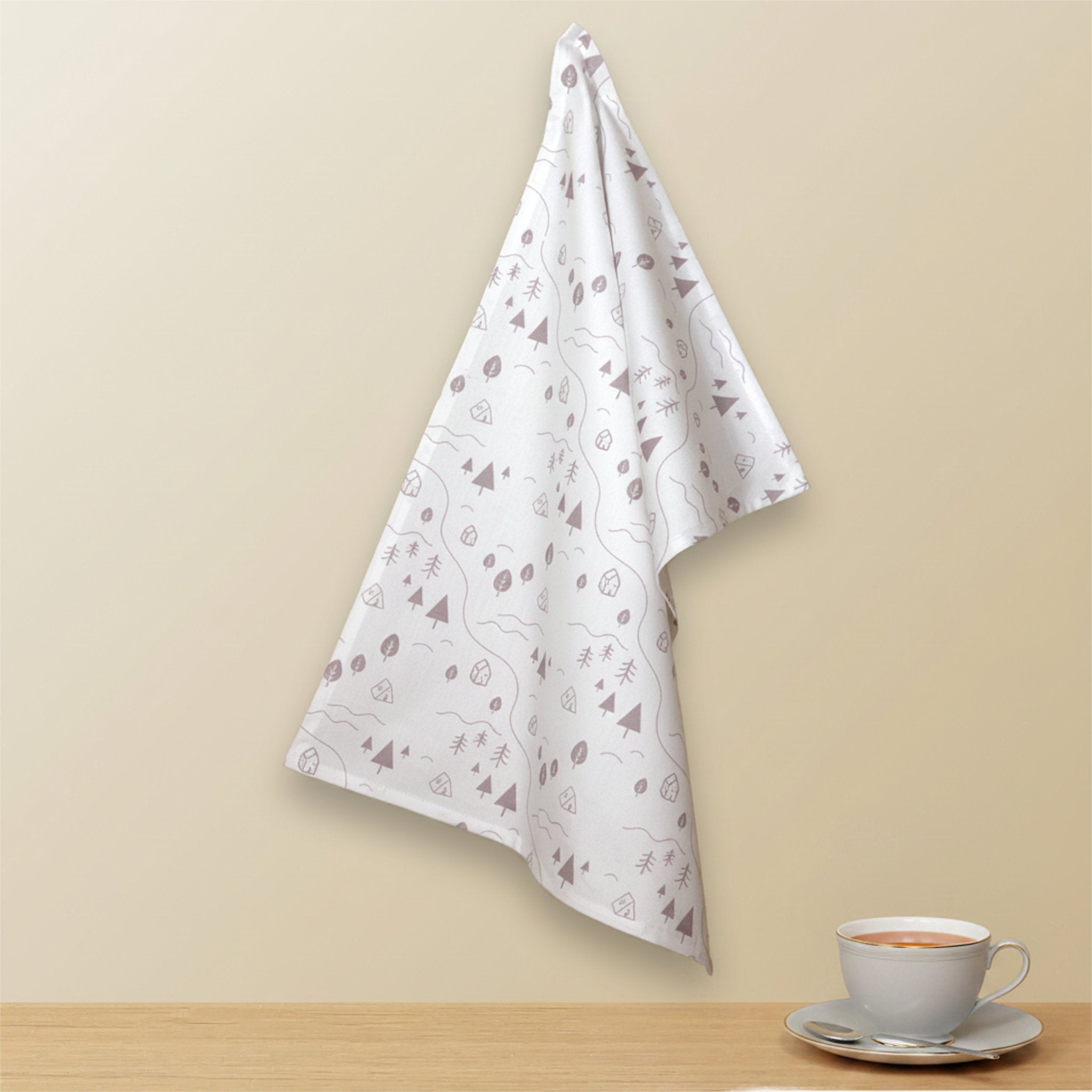 A white kitchen towel with a slate colored abstract pattern of tiny countryside icons hangs on a beige wall above a wooden table, with a white teacup and saucer on the right side from Hem And Tassel
