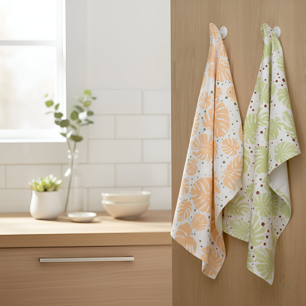 Two monstera leaf patterned kitchen towels hanging on a wooden cabinet with a potted plant on the kitchen counter. The towels are in peach, and light green on white backgrounds from Hem And Tassel 
