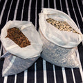 Two open organic cotton produce bags filled with seeds on a back and white striped background from Hem And Tassel 